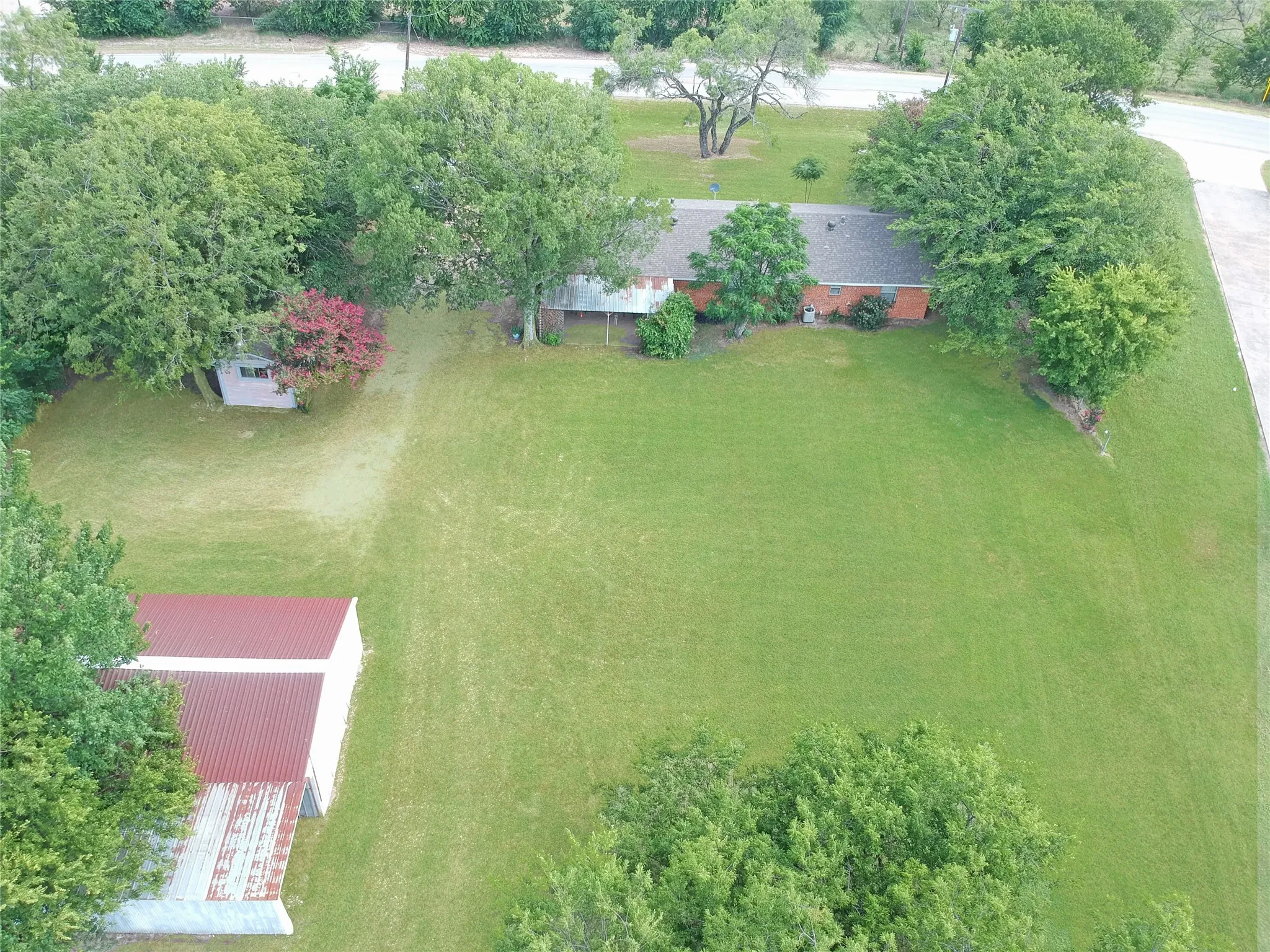 Expansive green lawn bordering a paved road, featuring a single-story residence with a dark roof and brick exterior, mature trees, and a separate structure with a red metal roof
