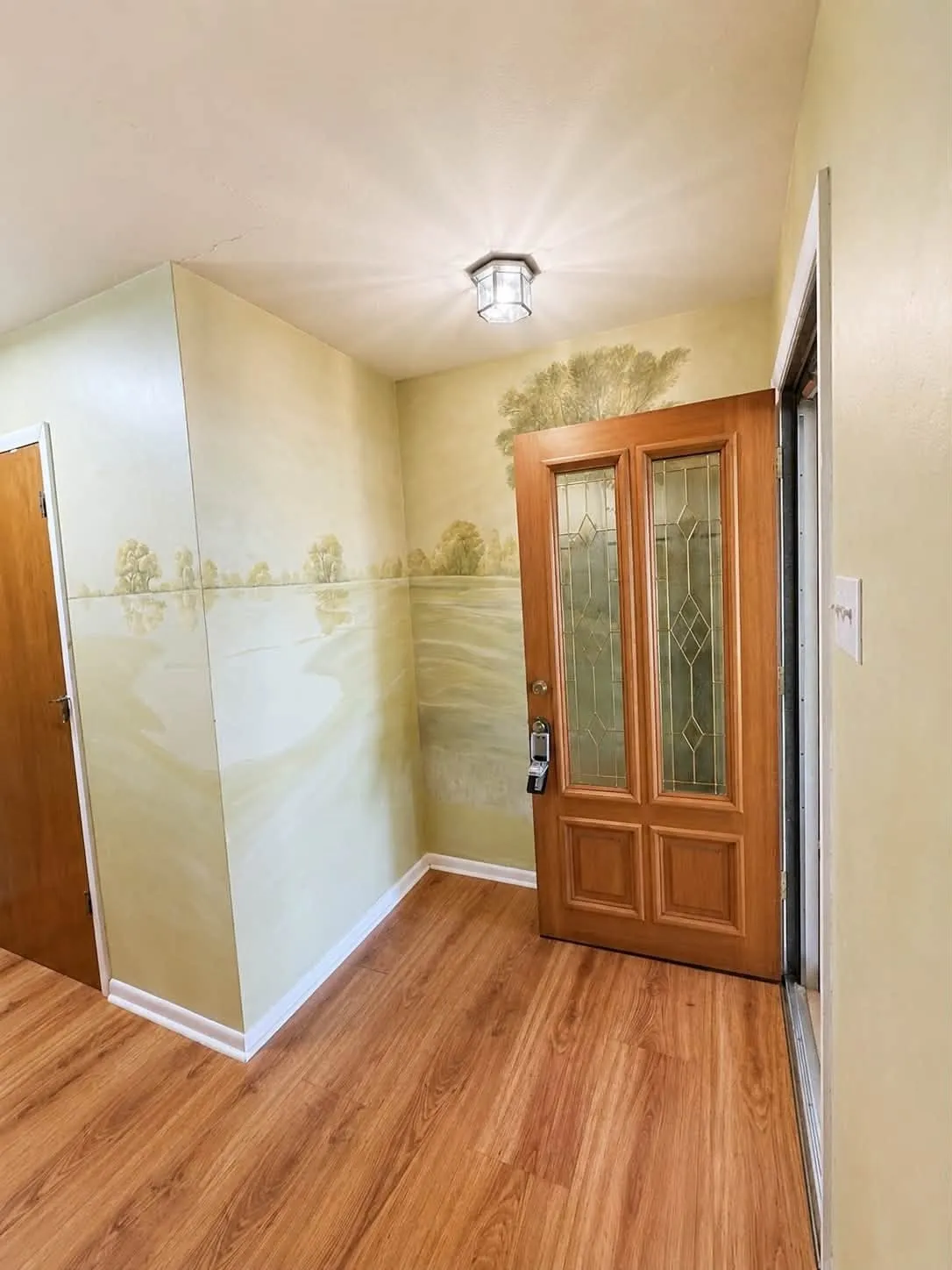 Foyer featuring a wood-finish floor, detailed wall mural, flush-mount ceiling light, and a wooden entry door with decorative glass panels