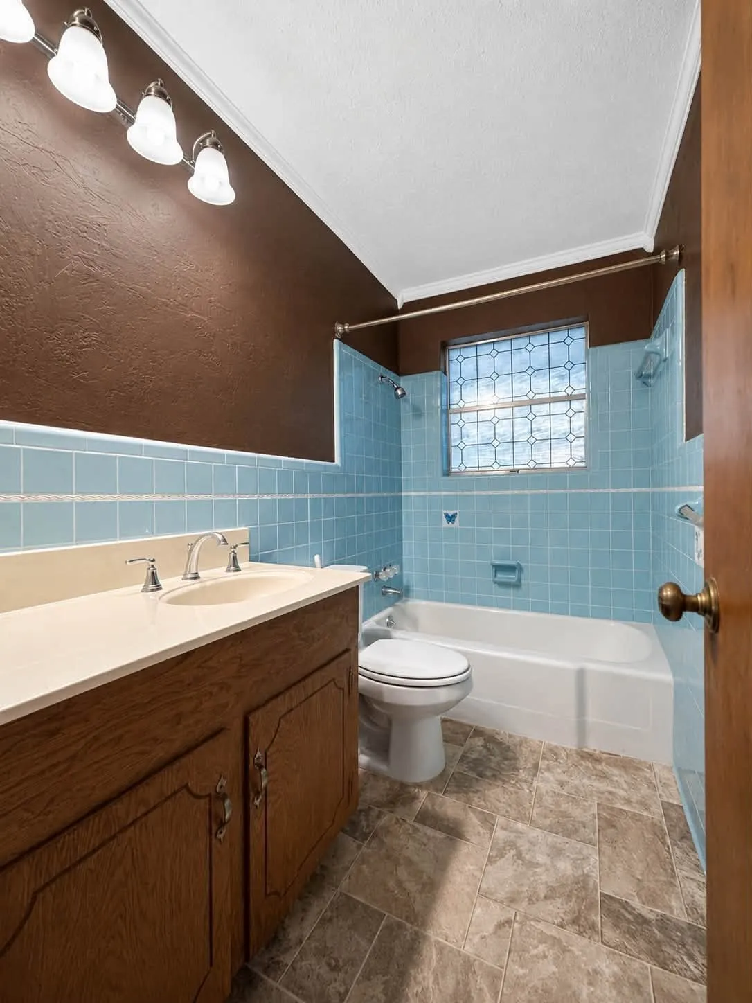 Bathroom featuring a wood-finish vanity with a light-toned countertop, a full bathtub with blue tile surround, a toilet, and patterned floor tiling