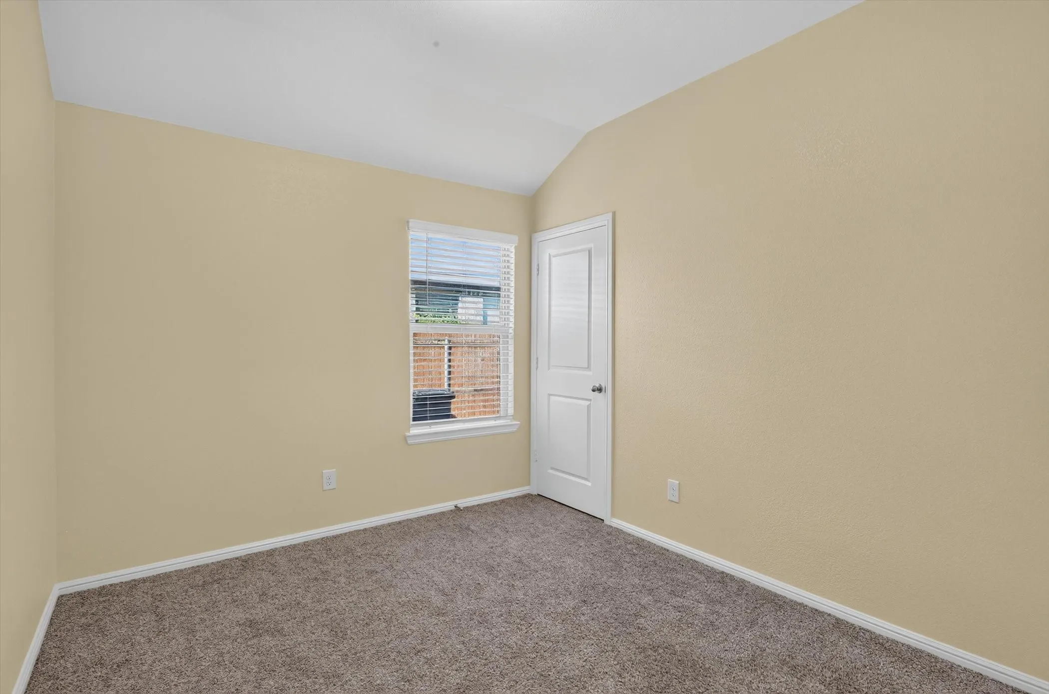 Carpeted room with a vaulted ceiling, a single window with blinds, and a white paneled door