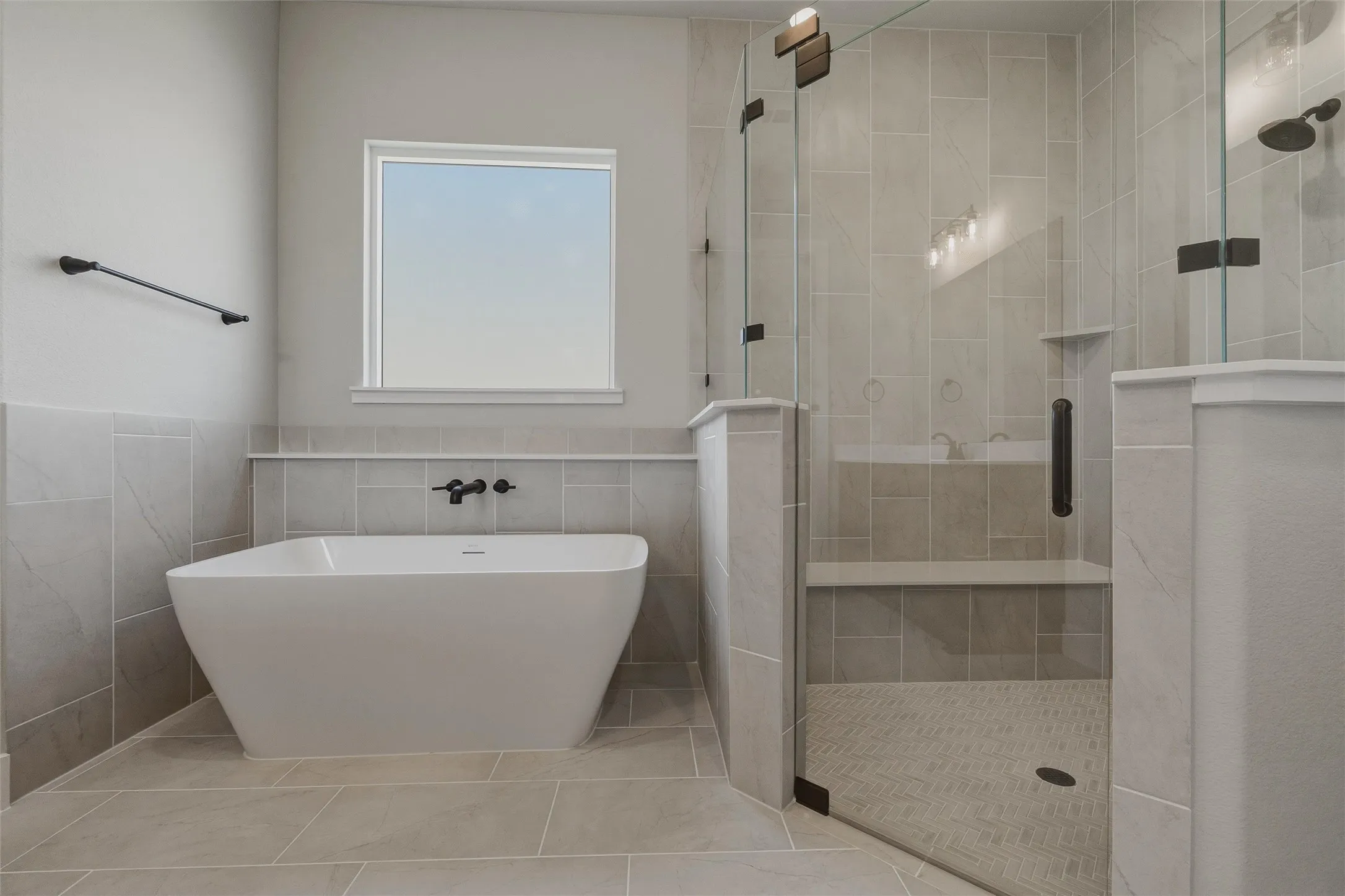 Freestanding soaking tub with wall-mounted matte black fixtures, glass-enclosed shower with a built-in bench, and large format tile flooring