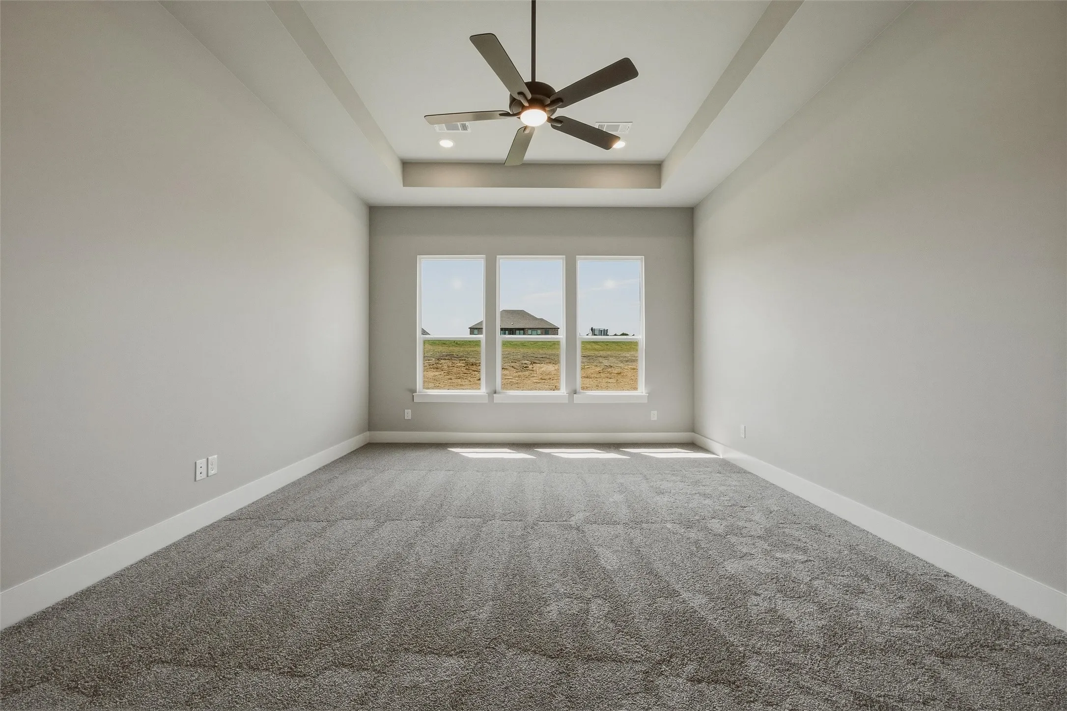 Tray ceiling with recessed lighting and a ceiling fan, three large windows, gray carpeting, and light gray wall finishes