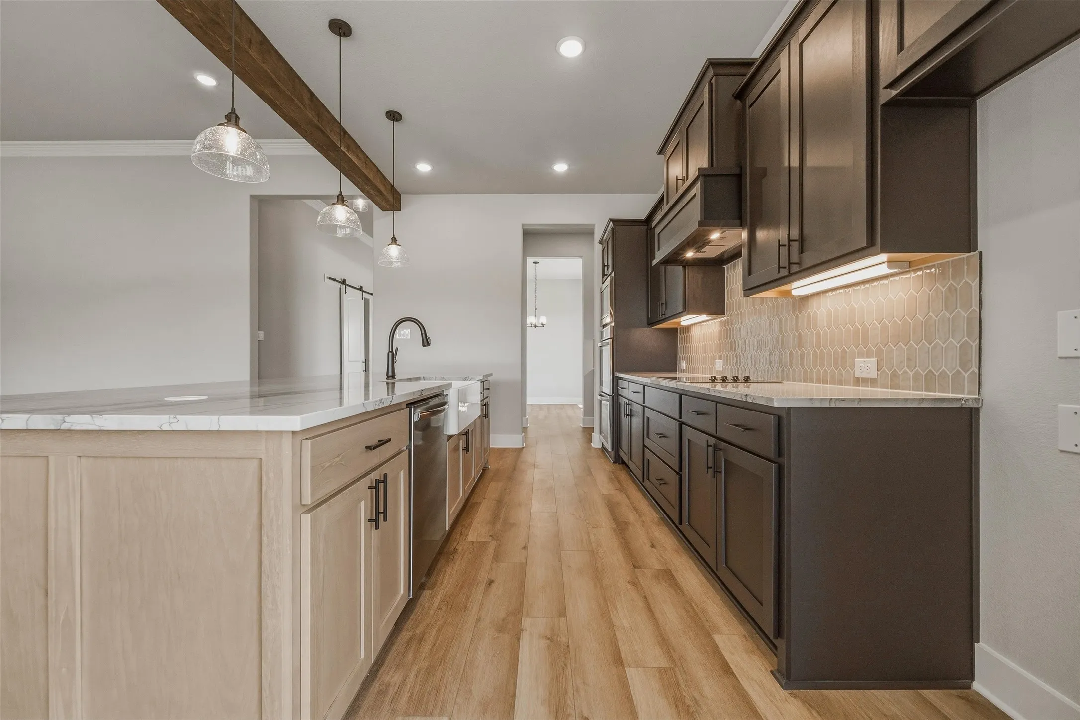 Kitchen featuring wood-finish flooring, an island with light-toned cabinetry and a farmhouse sink, dark-toned perimeter cabinetry, under-cabinet lighting, and a geometric tile backsplash