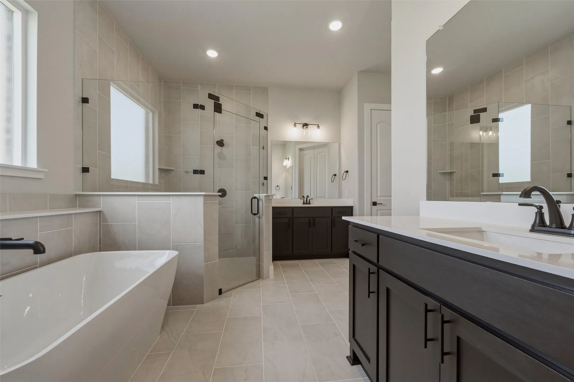 Freestanding bathtub, glass-enclosed shower, dual vanities with dark wood-finish cabinetry, and neutral-toned tile flooring