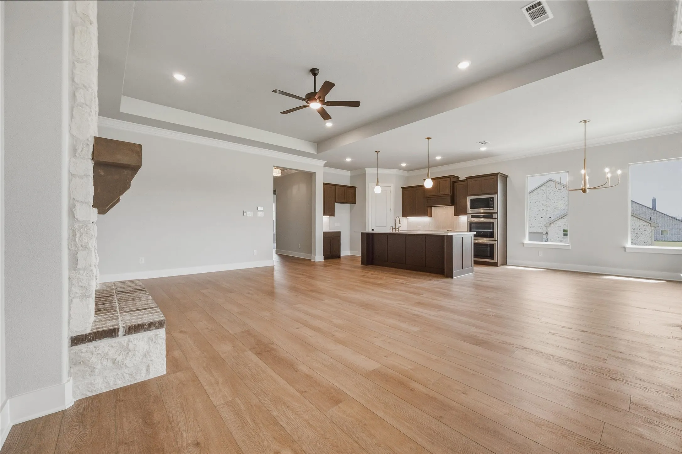 Wood-finish flooring throughout, tray ceiling with recessed lighting, stone-clad fireplace with wood mantel, contemporary ceiling fan, and a kitchen island with pendant lighting