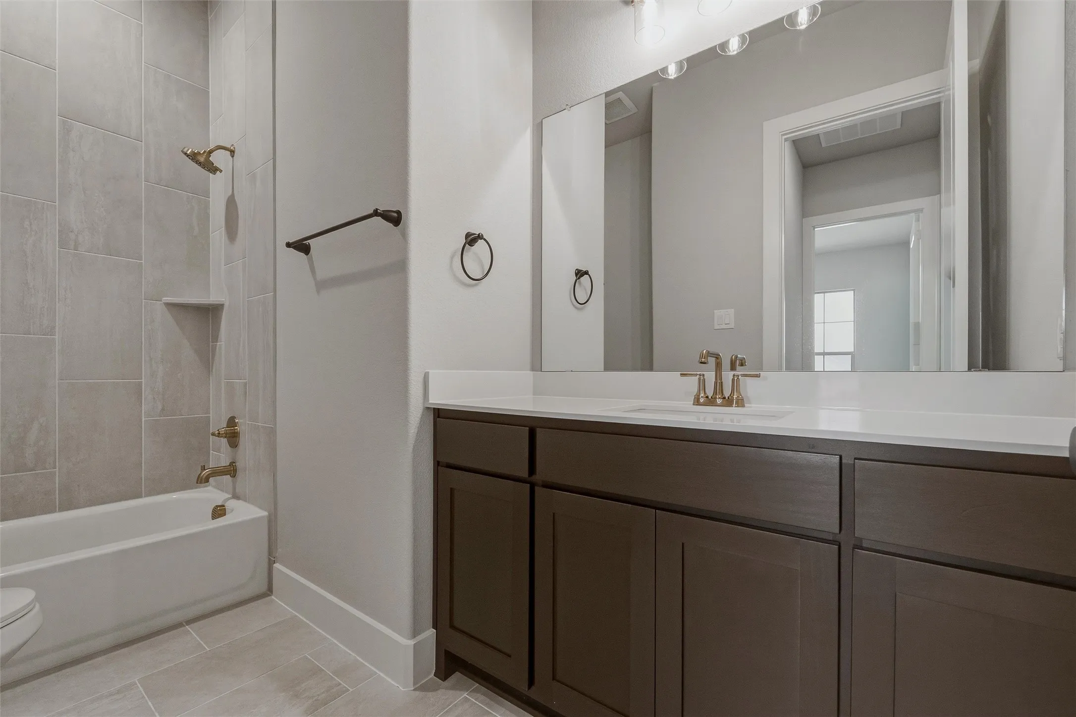 Bathroom featuring a large vanity with a white countertop and dark cabinetry, a spacious mirror, and a bathtub with tile surround and brass fixtures