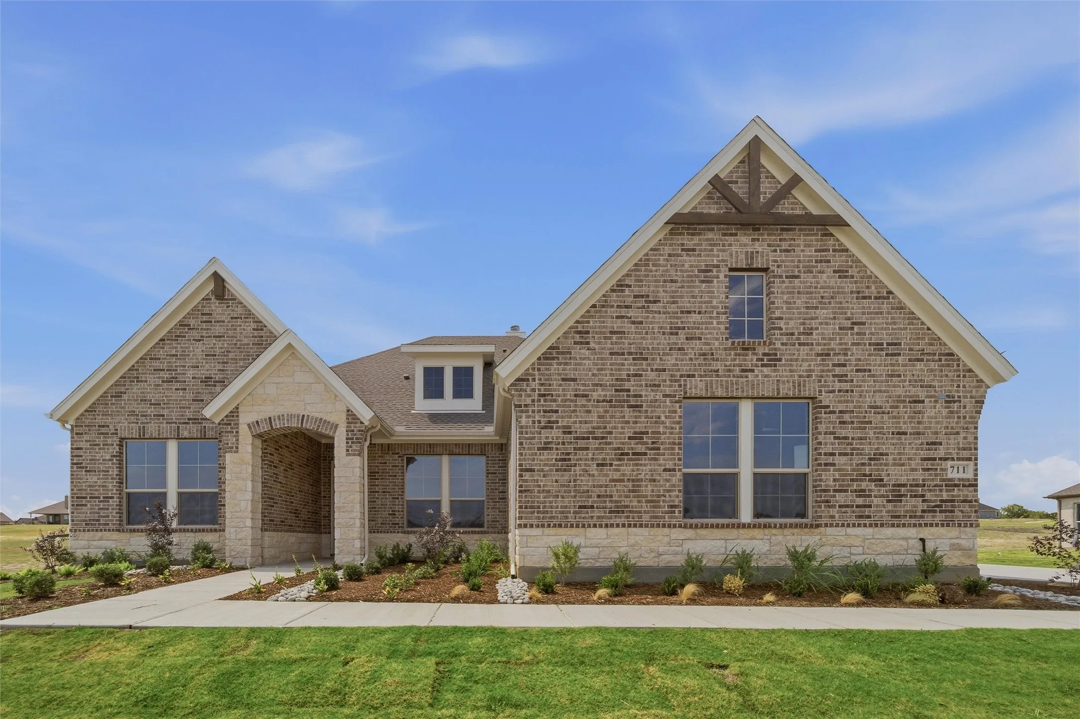 Brick and stone facade, gabled rooflines, decorative timber truss accent, established landscaping, and a concrete pathway