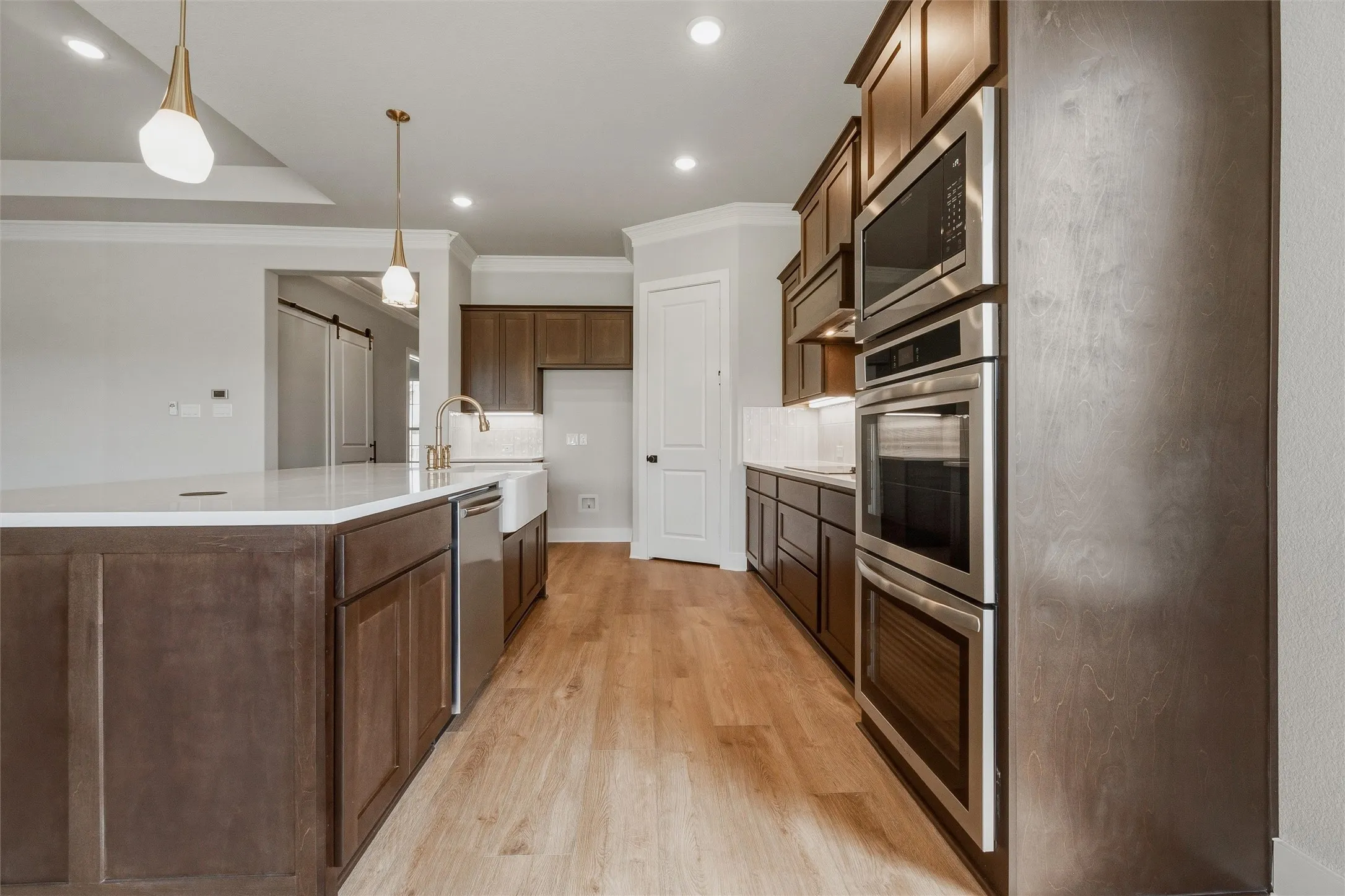 Kitchen featuring wood-finish flooring, dark wood cabinetry, stainless steel appliances, a white farmhouse sink with a gold-finish faucet, and pendant lighting