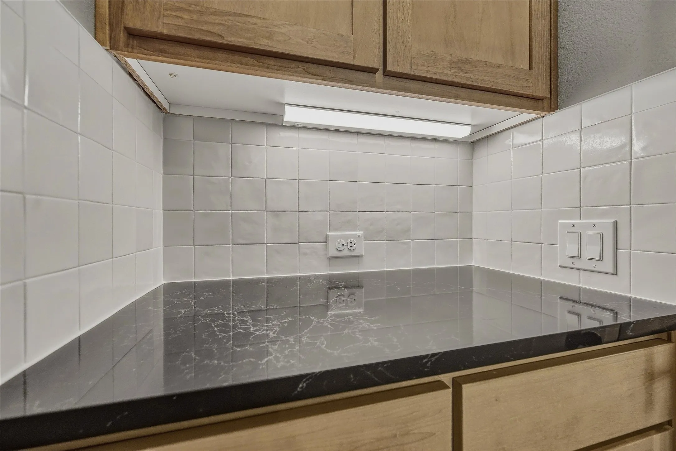 Under-cabinet lighting illuminating a dark countertop with light veining, subway tile backsplash, and wood-finish cabinetry