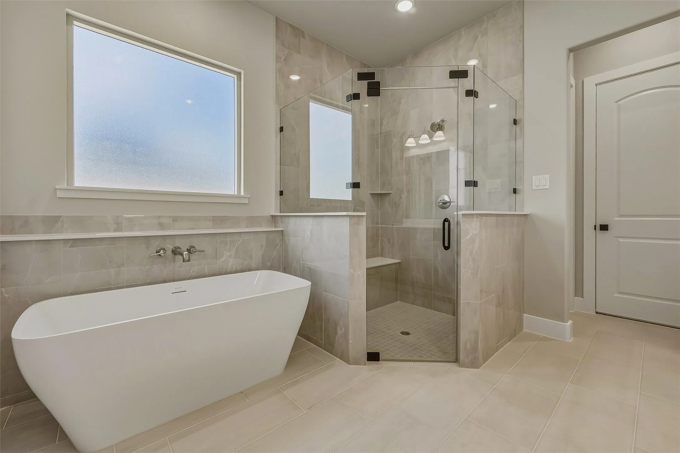 Freestanding soaking tub with a wall-mounted faucet, walk-in shower featuring a built-in bench, light-toned tile flooring, and a privacy window