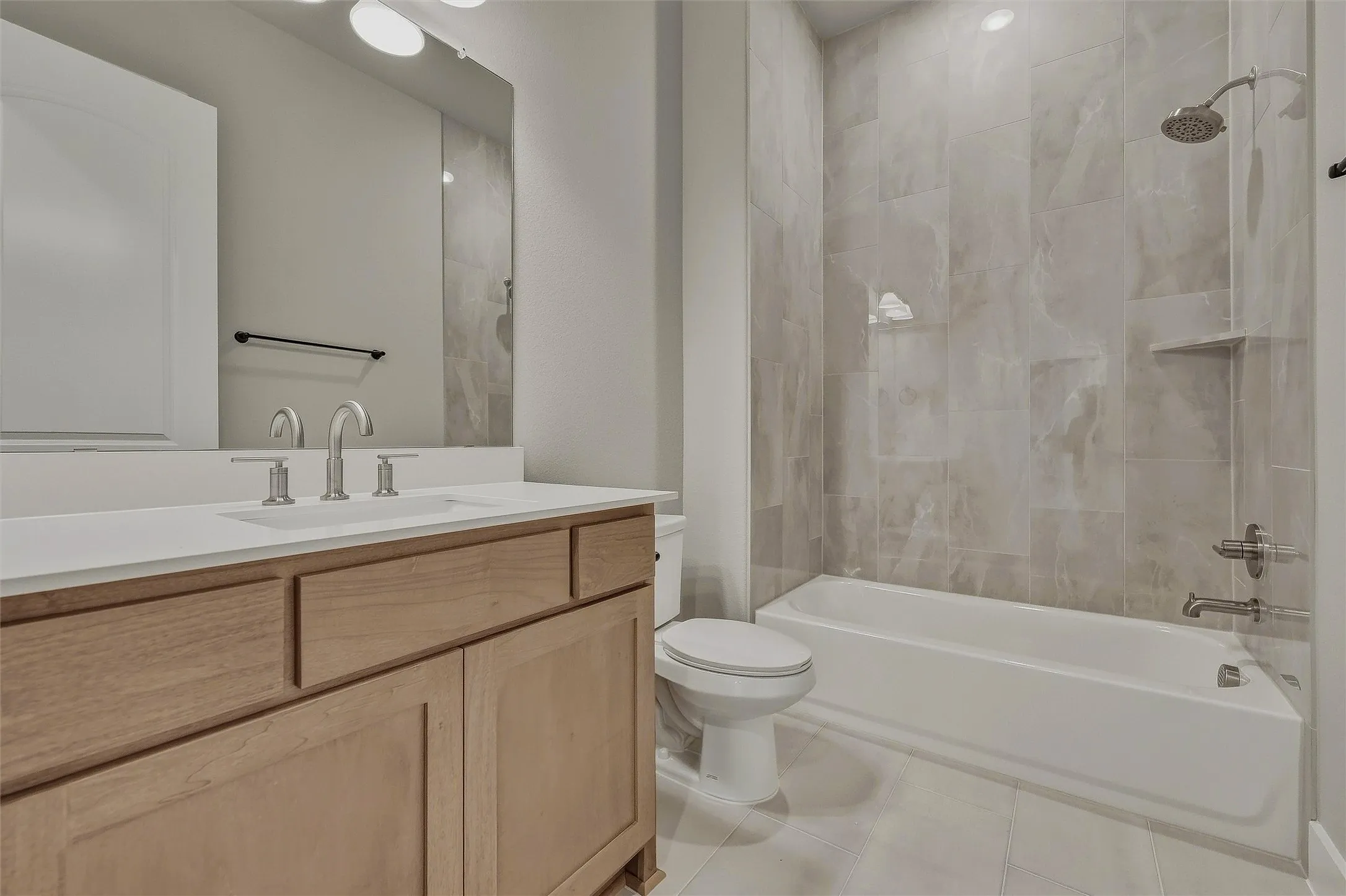 Spacious bathroom featuring a single vanity with light wood cabinetry, a large vanity mirror, a drop-in sink, brushed nickel fixtures, and a shower-tub combination with tile surround