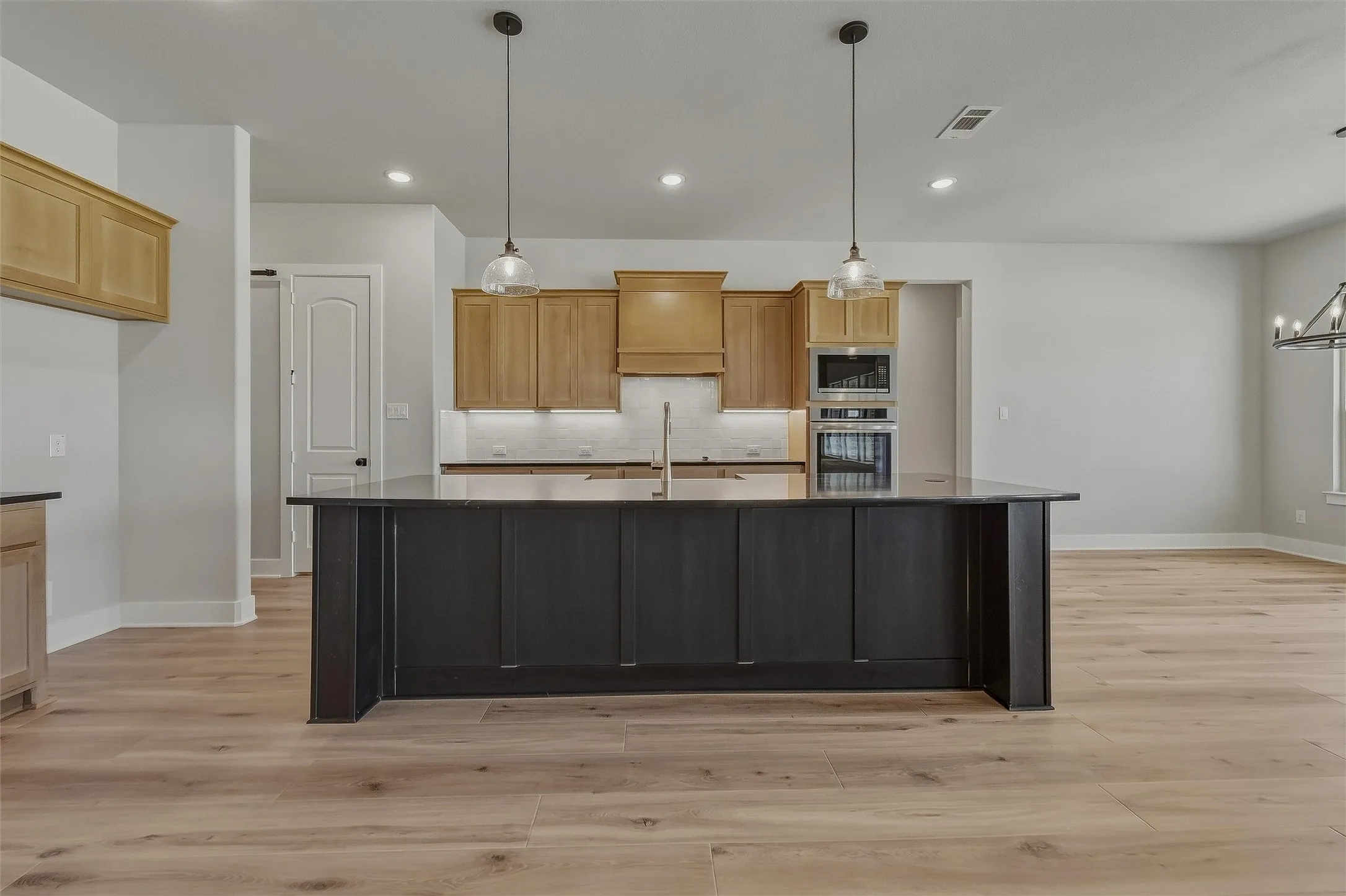 Spacious kitchen featuring a dark-toned island with ample countertop space, light wood-finish cabinetry, stainless steel double ovens, and wood-finish flooring throughout