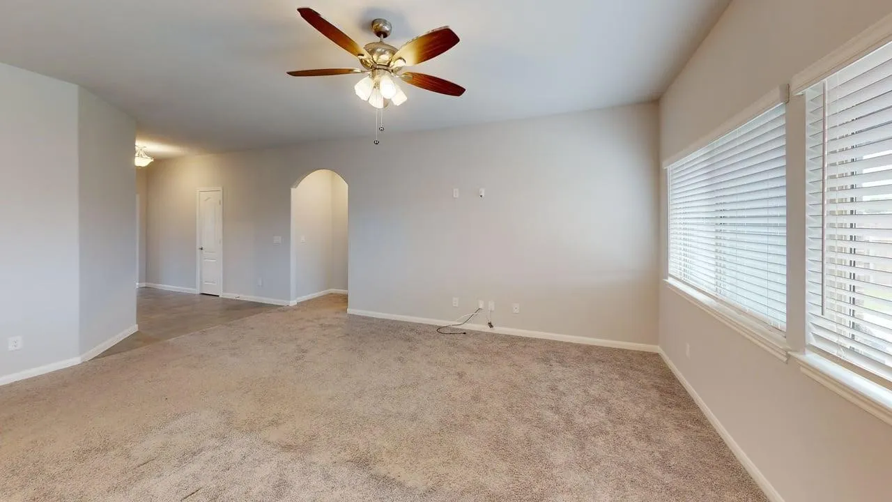 Expansive carpeted room featuring a ceiling fan with integrated lighting, two windows with blinds, and an arched doorway