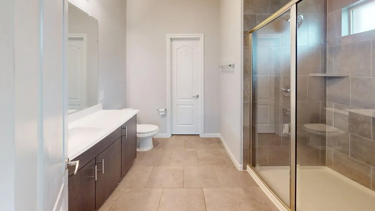 Bathroom featuring a glass-enclosed shower with tile surround, a built-in shower niche, a long vanity with dual sinks and dark wood-finish cabinetry, and neutral-toned tile flooring