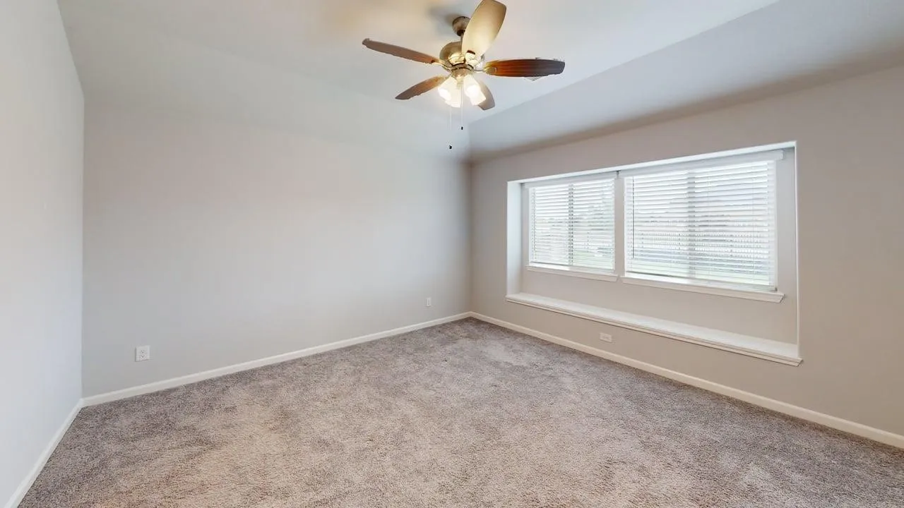 Spacious interior room featuring a tray ceiling with a ceiling fan, plush carpet flooring, and a wide window with blinds