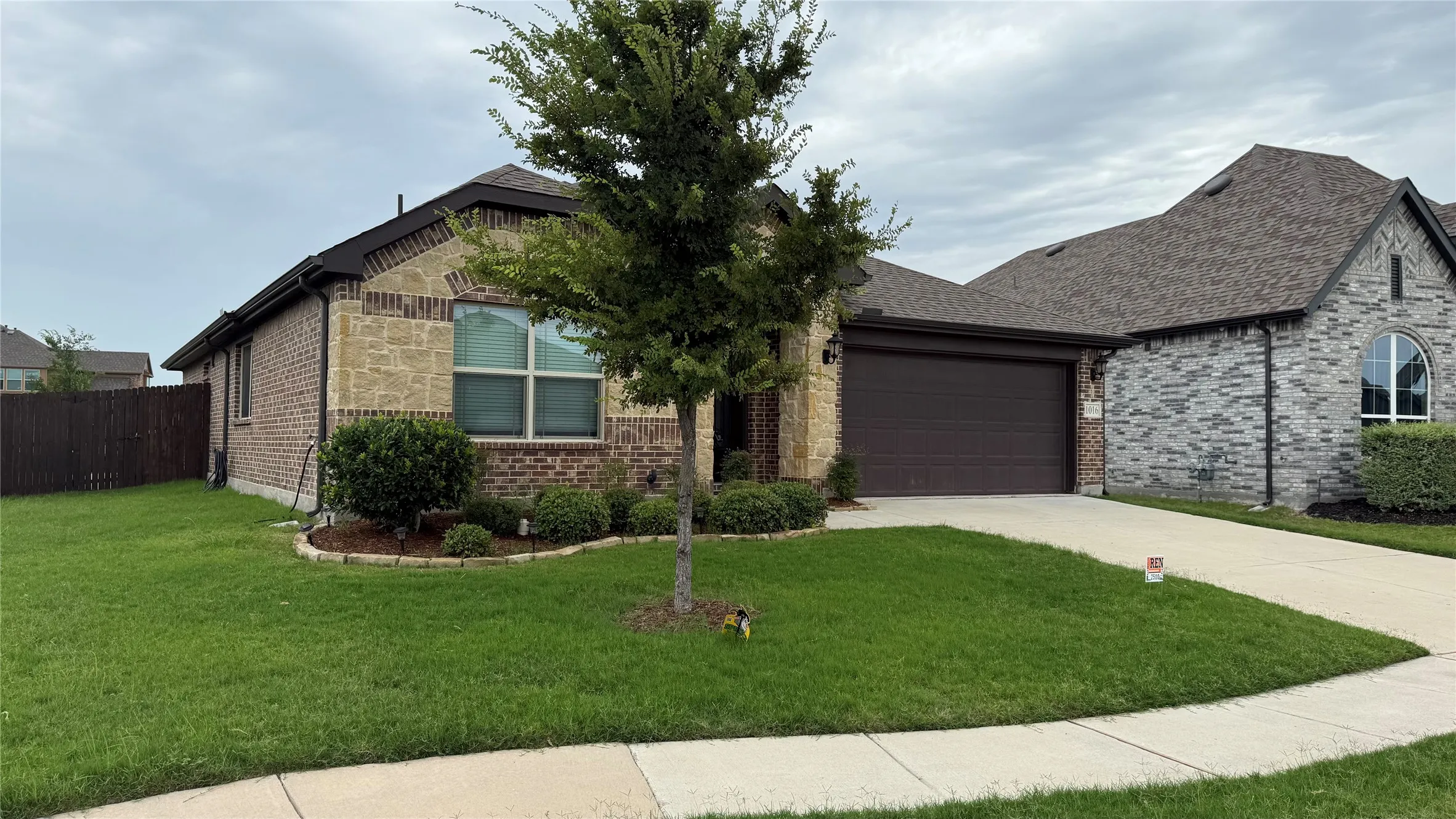 Brick and stone facade residence featuring a dark brown garage door, shingle roof, and a mature tree in the front yard