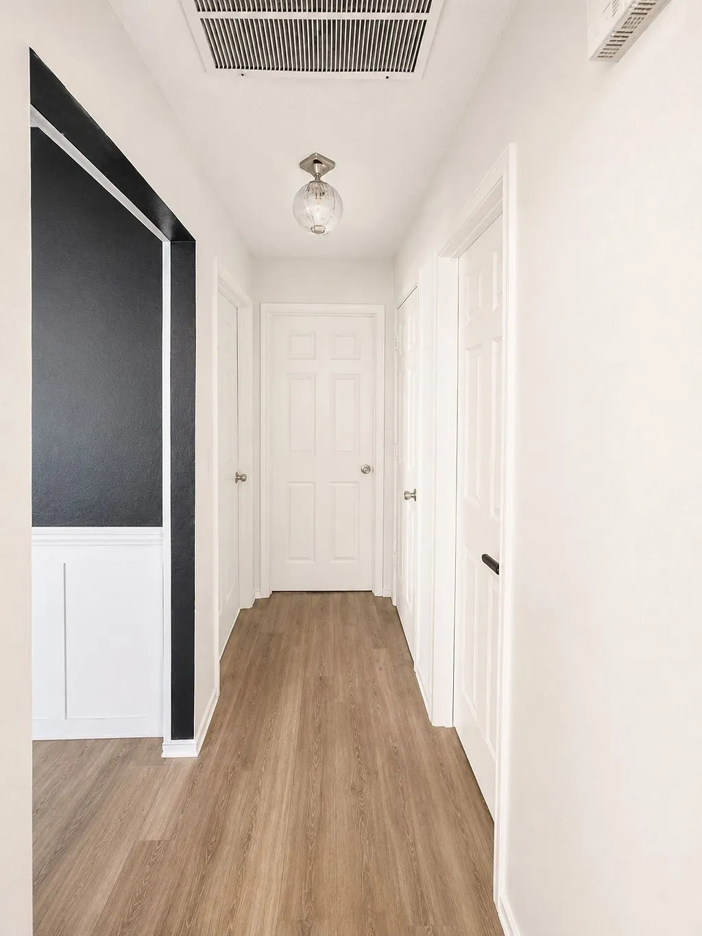 Hallway featuring wood-finish flooring, white paneled doors, wainscoting with a dark accent wall, and a flush-mount ceiling light fixture