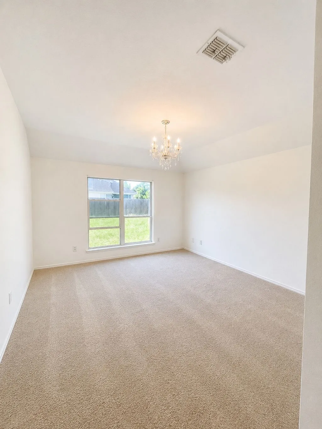 Spacious room featuring neutral carpeting, white painted walls, a crystal chandelier, and a large window overlooking a fenced yard