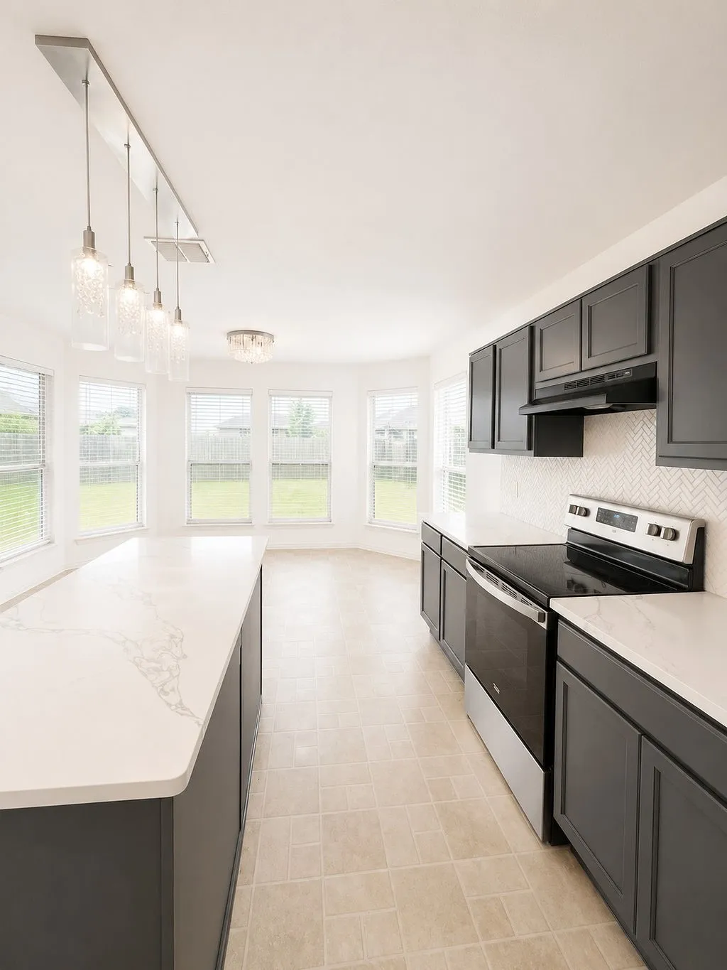 Spacious kitchen featuring an expansive island with a white veined countertop, pendant lighting, stainless steel appliances, dark cabinetry, and herringbone tile backsplash