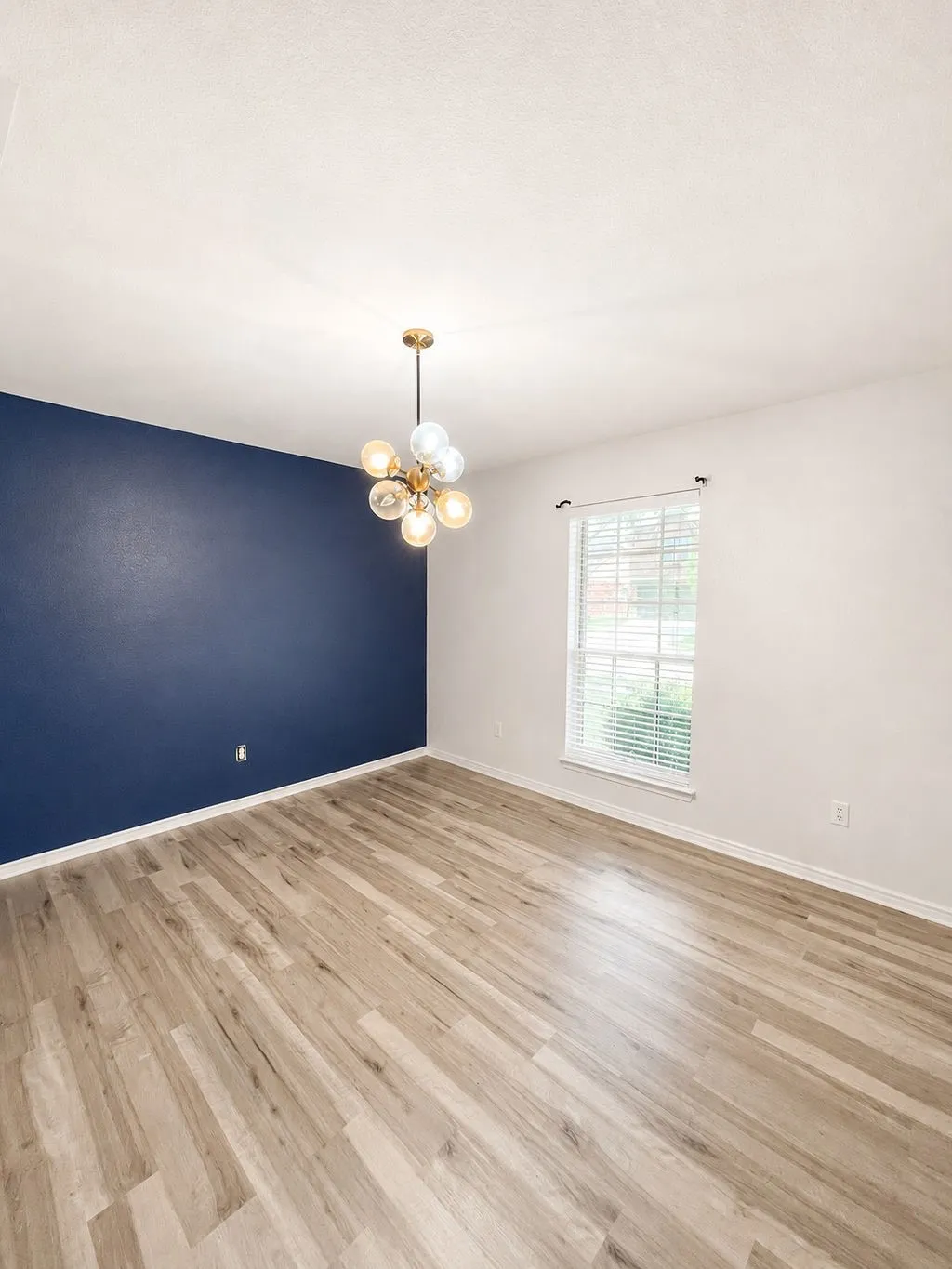 Spacious room featuring wood-finish flooring, a contemporary gold-tone chandelier with clear and frosted globes, and a prominent accent wall