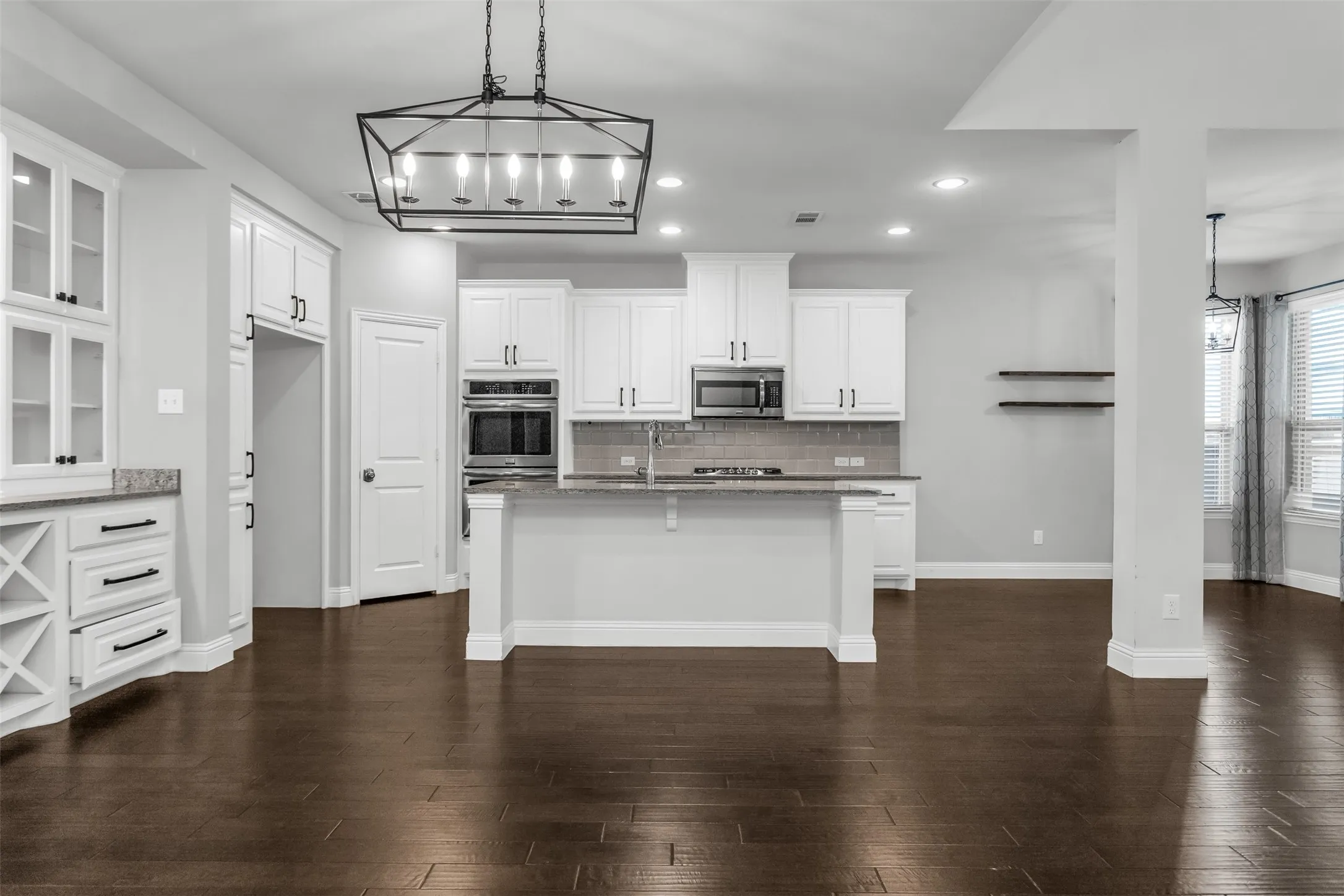 Spacious kitchen featuring an island with counter seating, a built-in oven and microwave, dark wood-finish flooring, white cabinetry with black hardware, and an overhead geometric chandelier