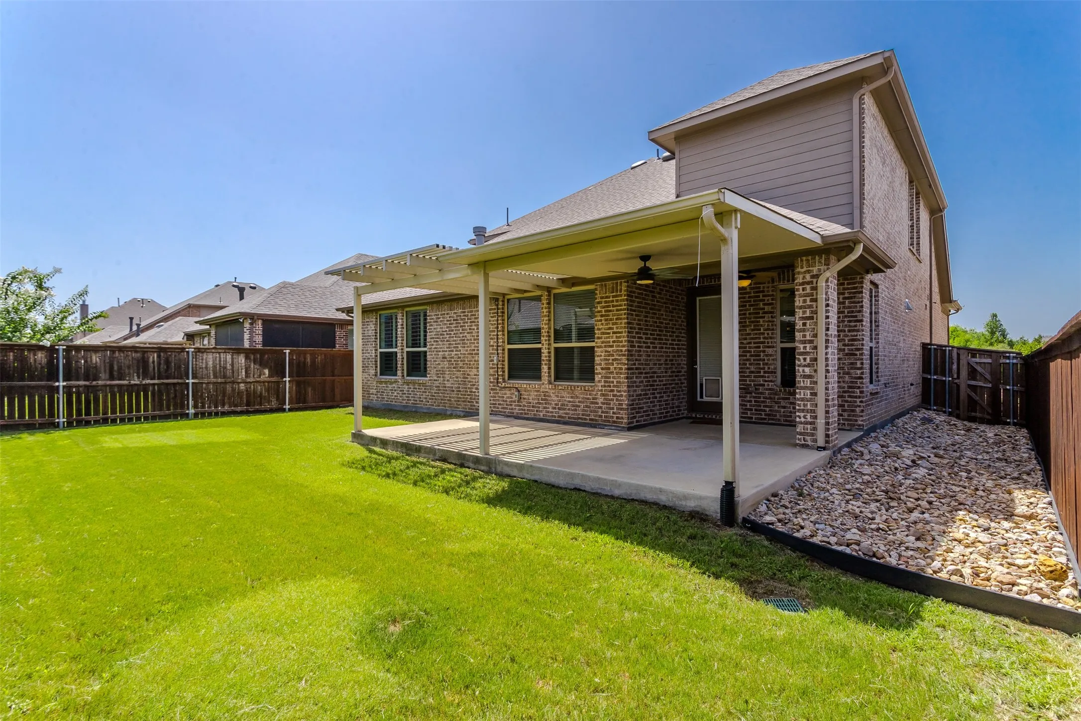 Expansive backyard featuring a covered patio with a ceiling fan, a lush green lawn, a wooden privacy fence, and a rock-filled drainage area along the side of the home