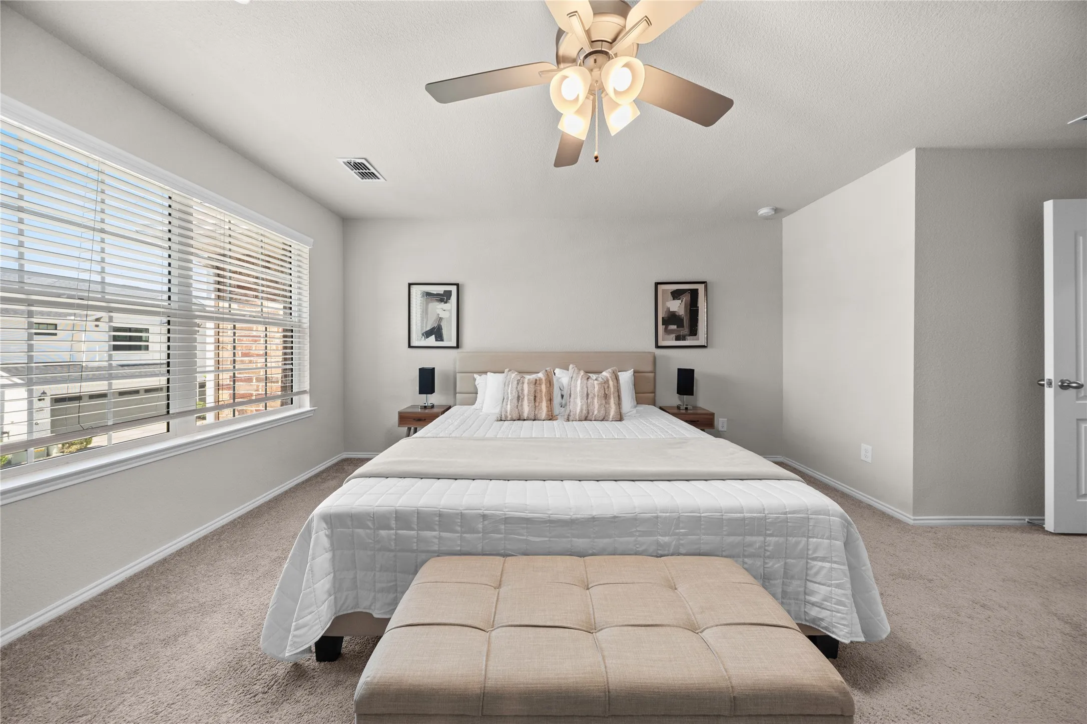 Expansive room featuring light gray walls, large window with blinds, ceiling fan, and neutral-toned carpeting