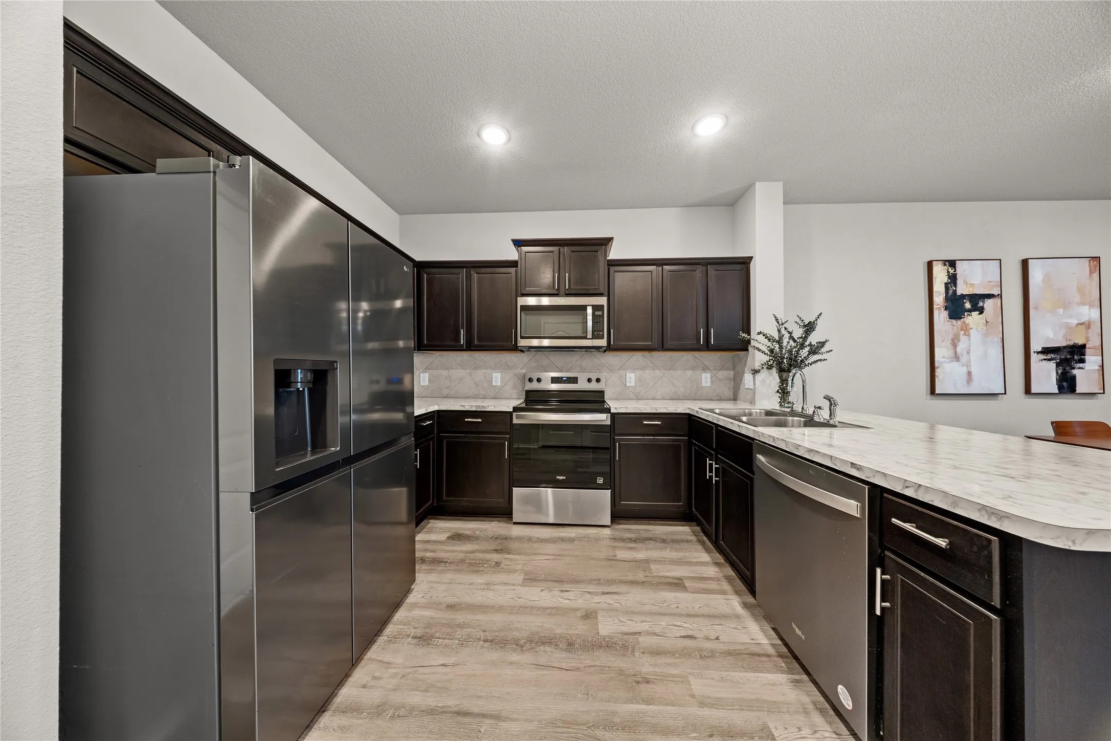 Modern kitchen featuring dark wood cabinetry, light wood-finish flooring, stainless steel appliances, a gray tiled backsplash, and a light-toned countertop