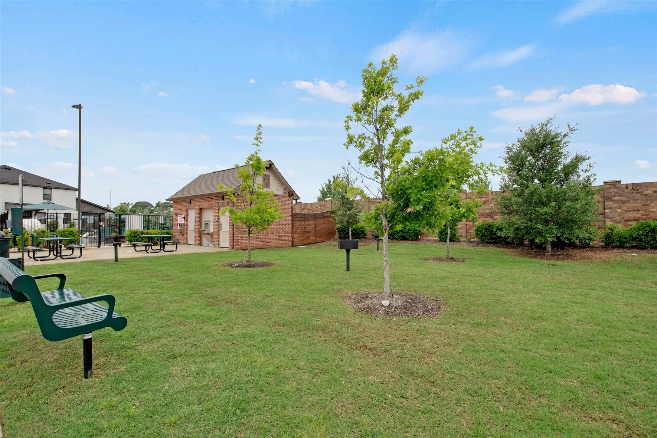 Expansive green space featuring a brick-clad amenity building, multiple picnic tables, a built-in outdoor grill, and mature trees