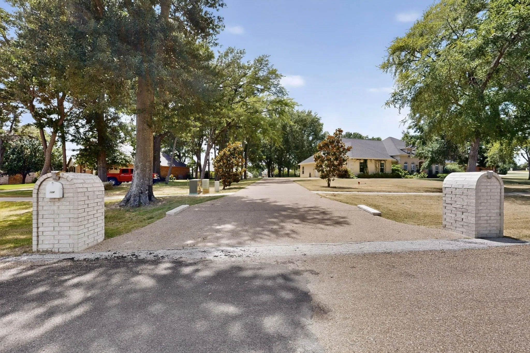Gravel driveway featuring brick mailbox pillars, mature trees, and a distant single-story residence with a dark roof