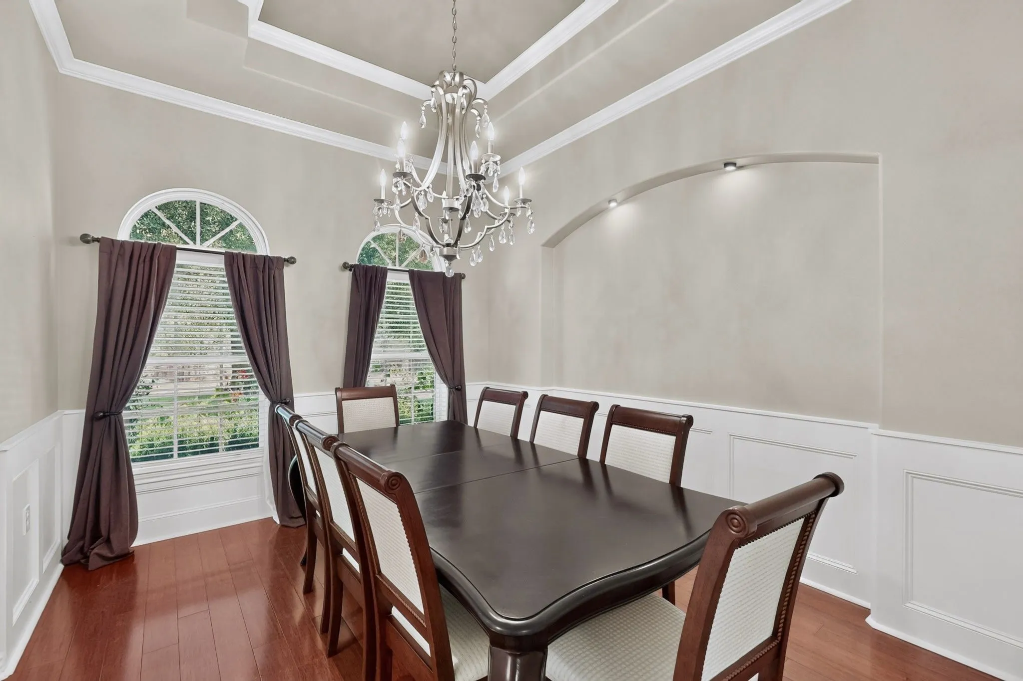 Formal dining area featuring hardwood flooring, wainscoting, an arched display niche with recessed lighting, a chandelier, and two arched windows
