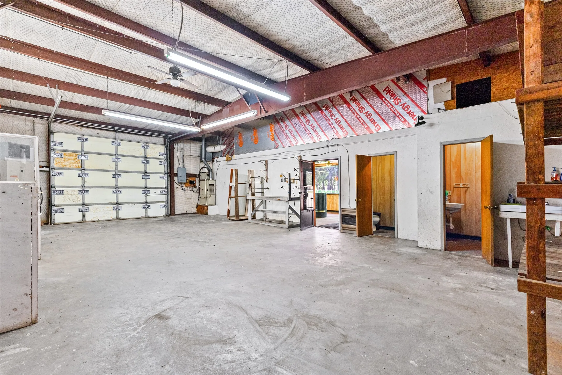 Spacious interior featuring a large roll-up door, exposed structural beams, high ceilings, multiple fluorescent lighting fixtures, and a ceiling fan
