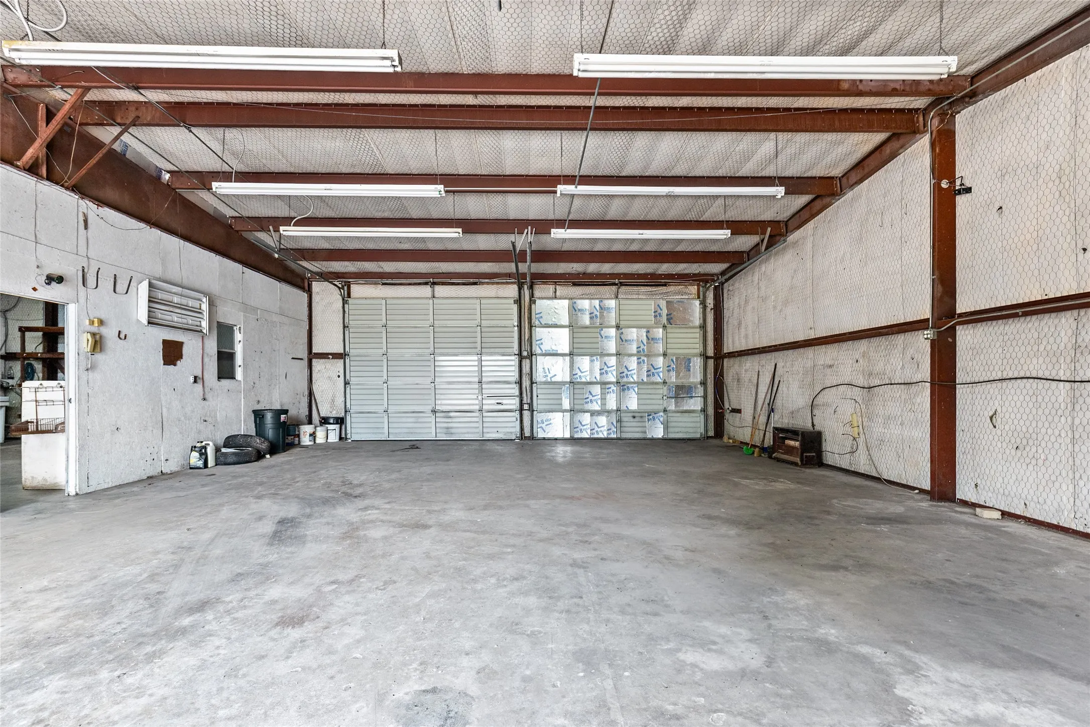 Spacious commercial interior featuring a concrete floor, exposed metal beam construction, multiple overhead fluorescent light fixtures, and two roll-up garage doors