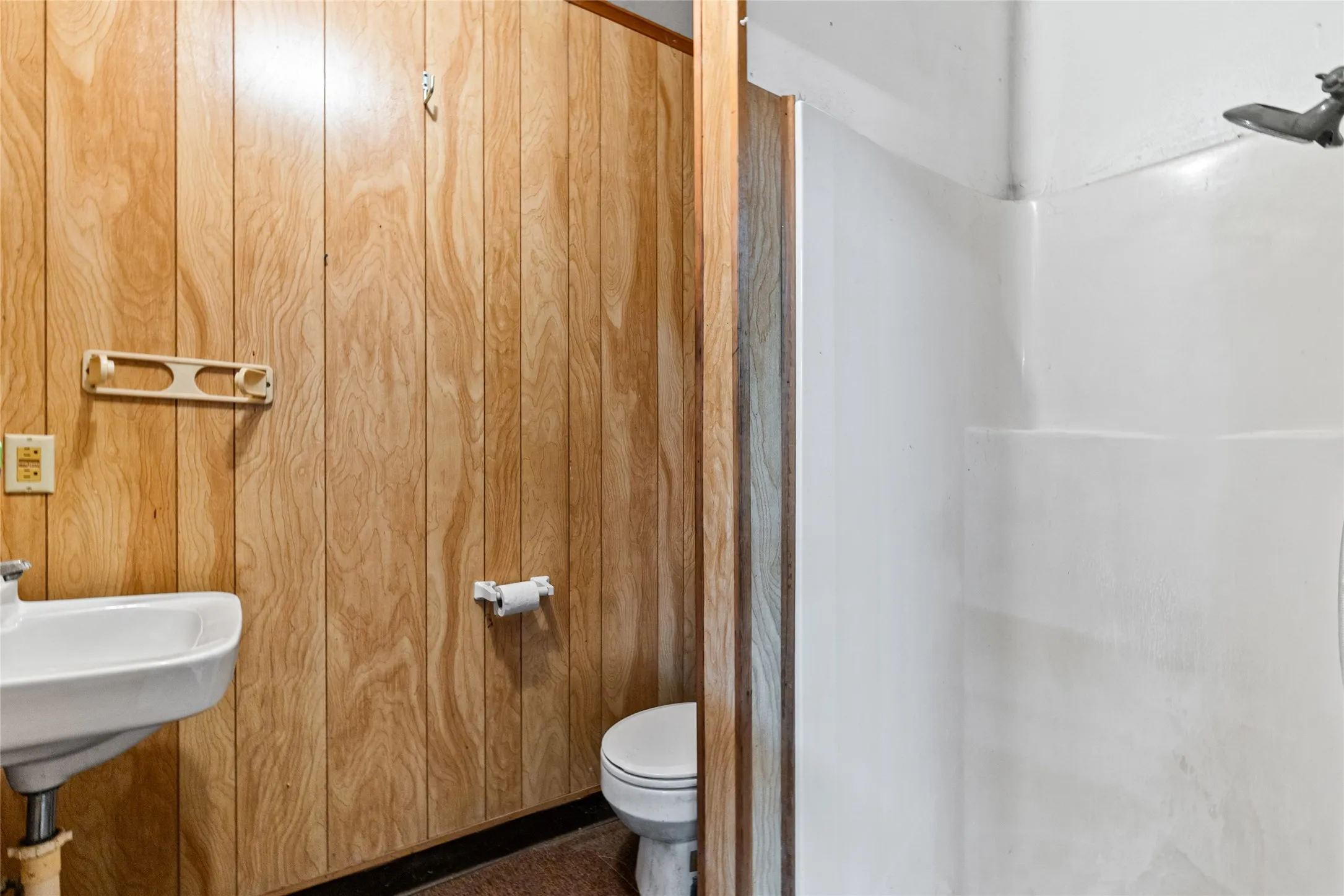 Wood-paneled bathroom featuring a white wall-mounted sink, toilet, and a white shower stall with a chrome showerhead