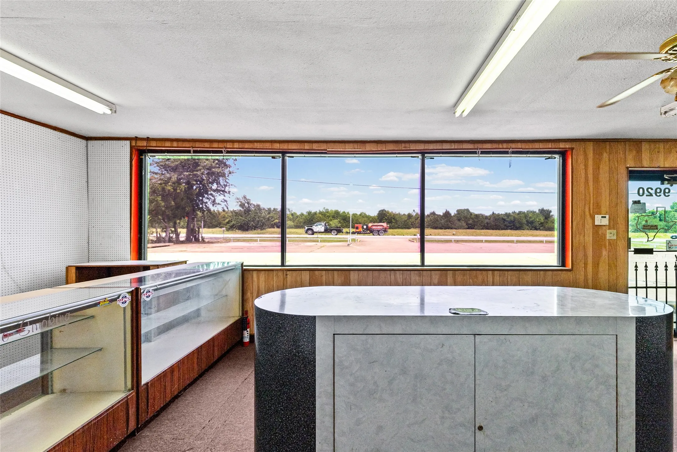 Expansive front window offering significant natural light, featuring a wood-paneled accent wall, track lighting, and a ceiling fan