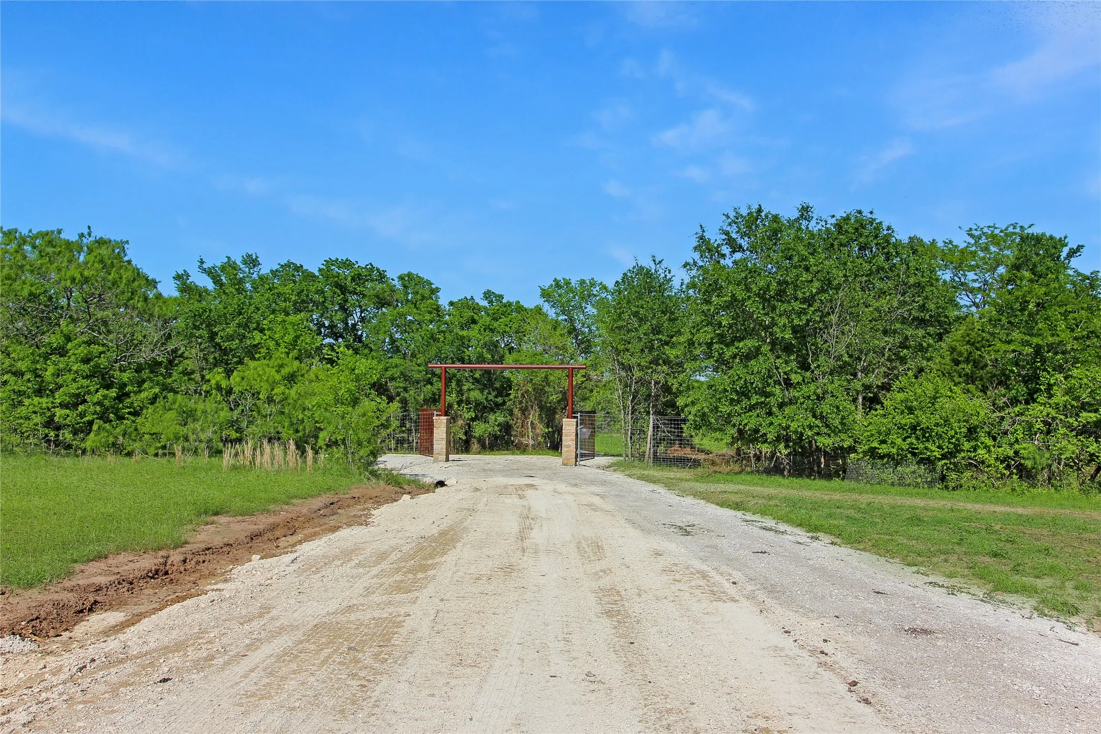 Gravel driveway leading to a metal gate with stone pillars, bordered by mature trees and natural landscaping