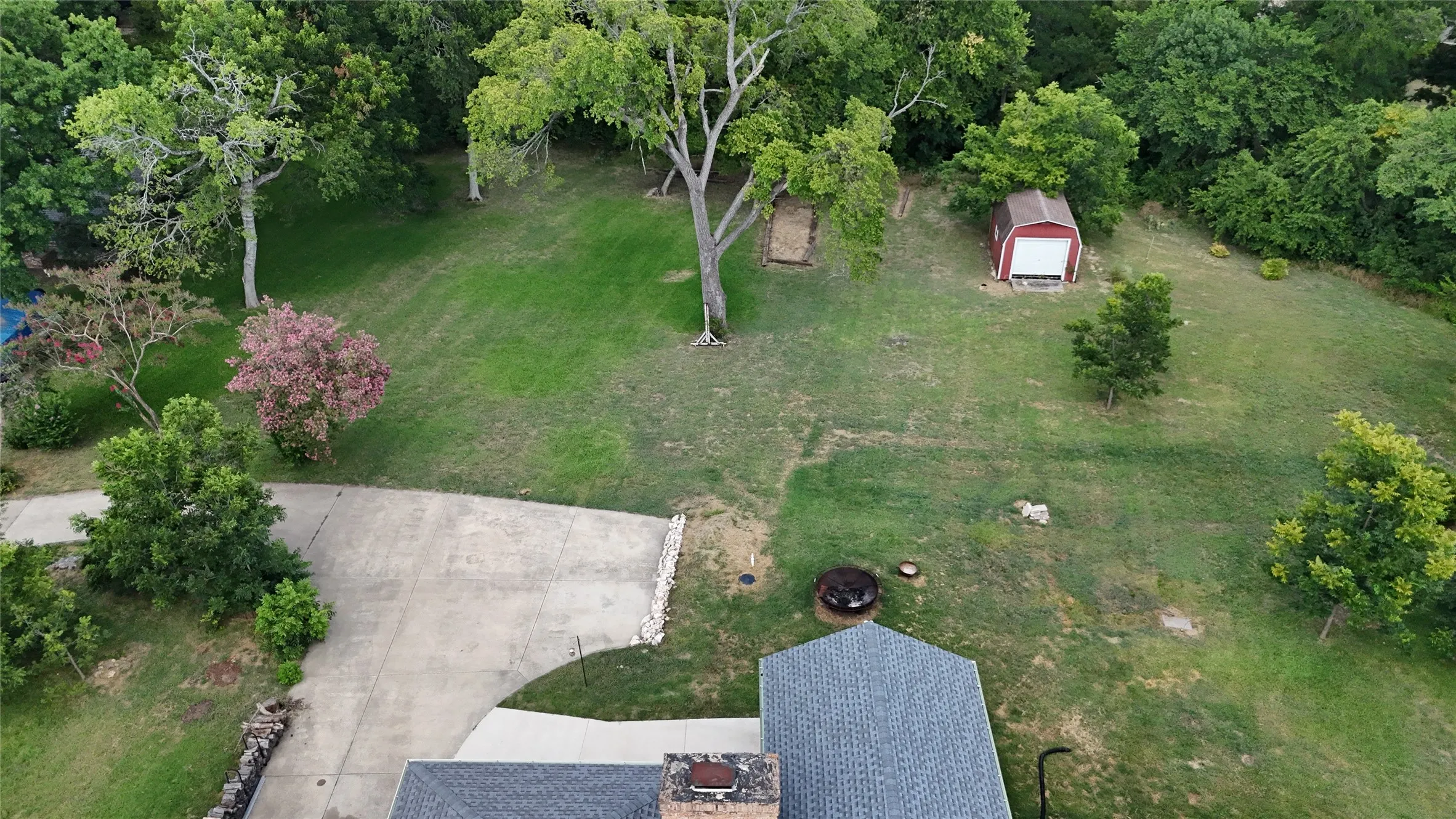 Expansive property featuring a paved driveway, mature trees, a red storage shed, and a fire pit area