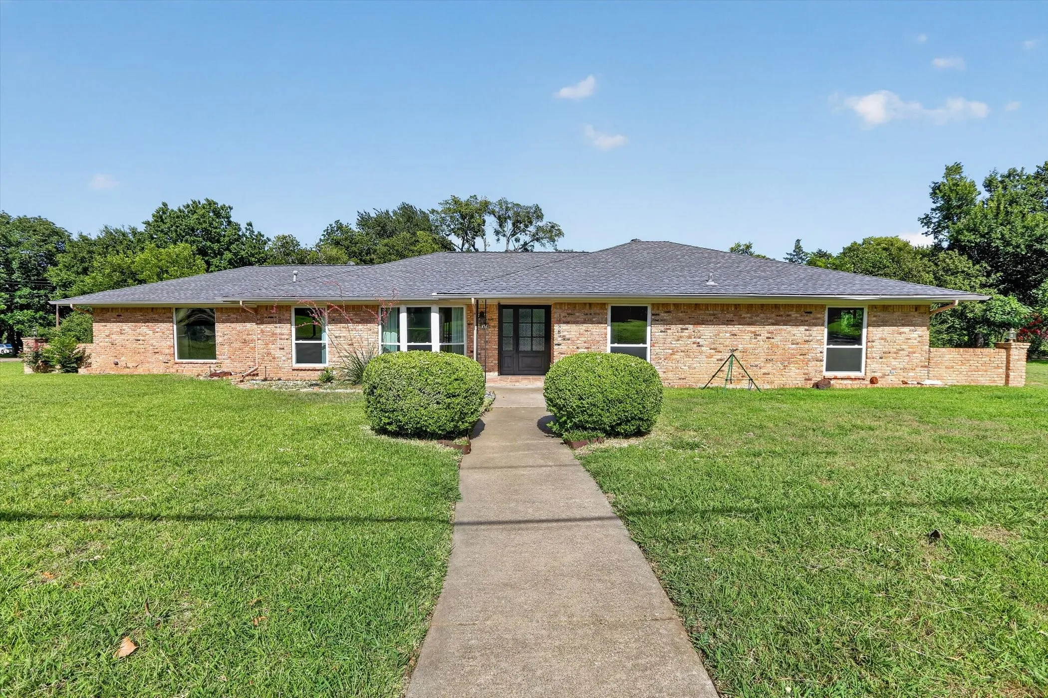 Brick exterior residence featuring a dark shingle roof and a concrete walkway flanked by manicured shrubbery