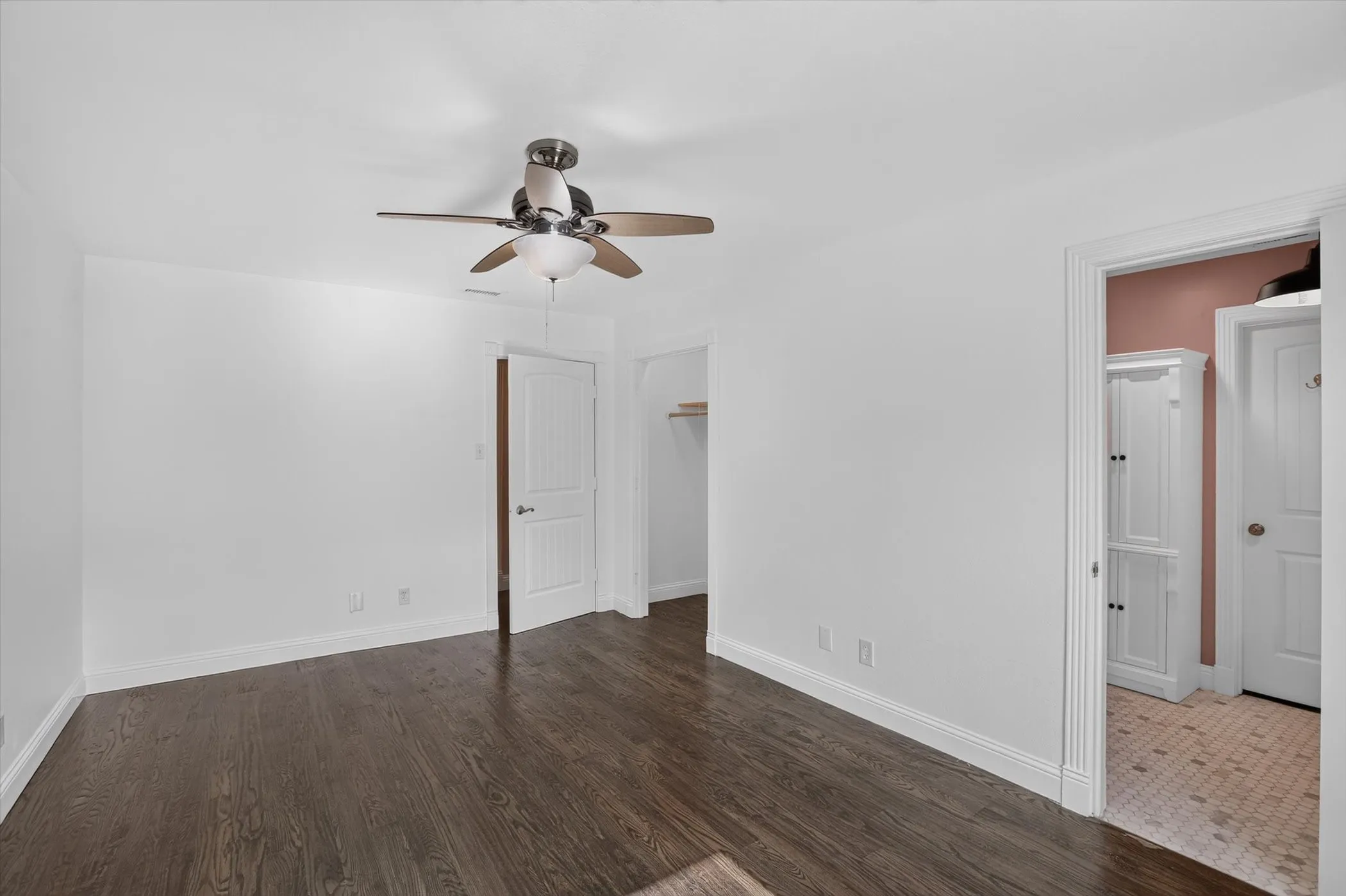 Spacious room featuring dark wood-finish flooring, white walls, and a ceiling fan with integrated lighting