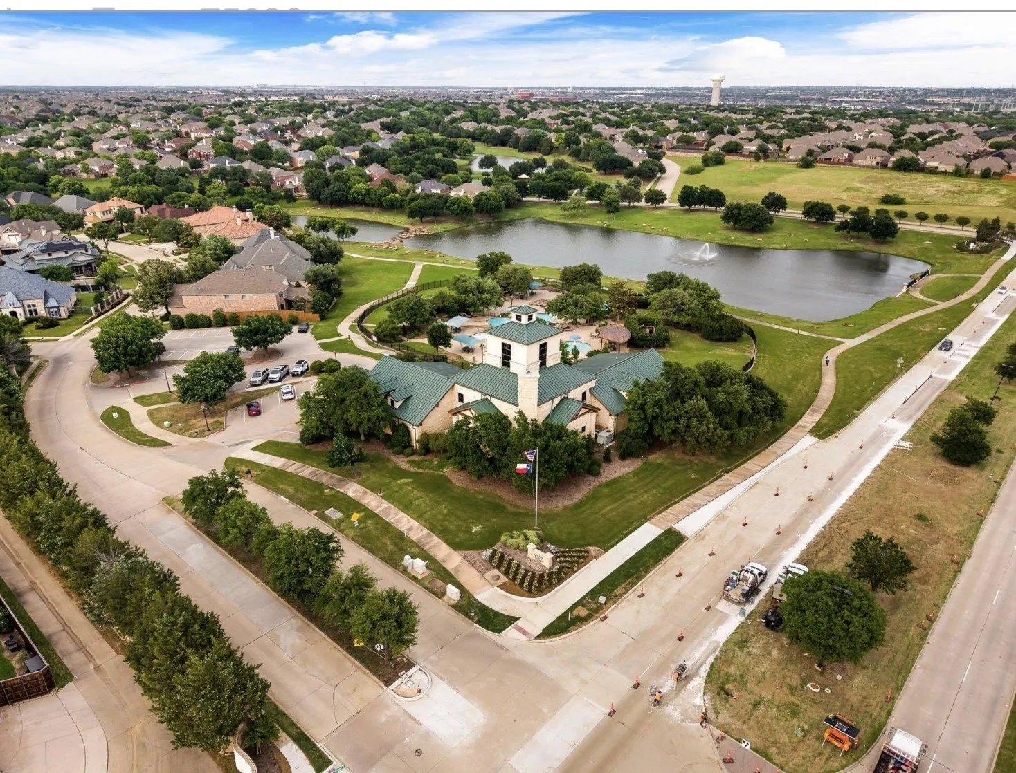 Expansive community grounds featuring a central building with green roofing, a large pond with a fountain, pedestrian pathways, and mature landscaping
