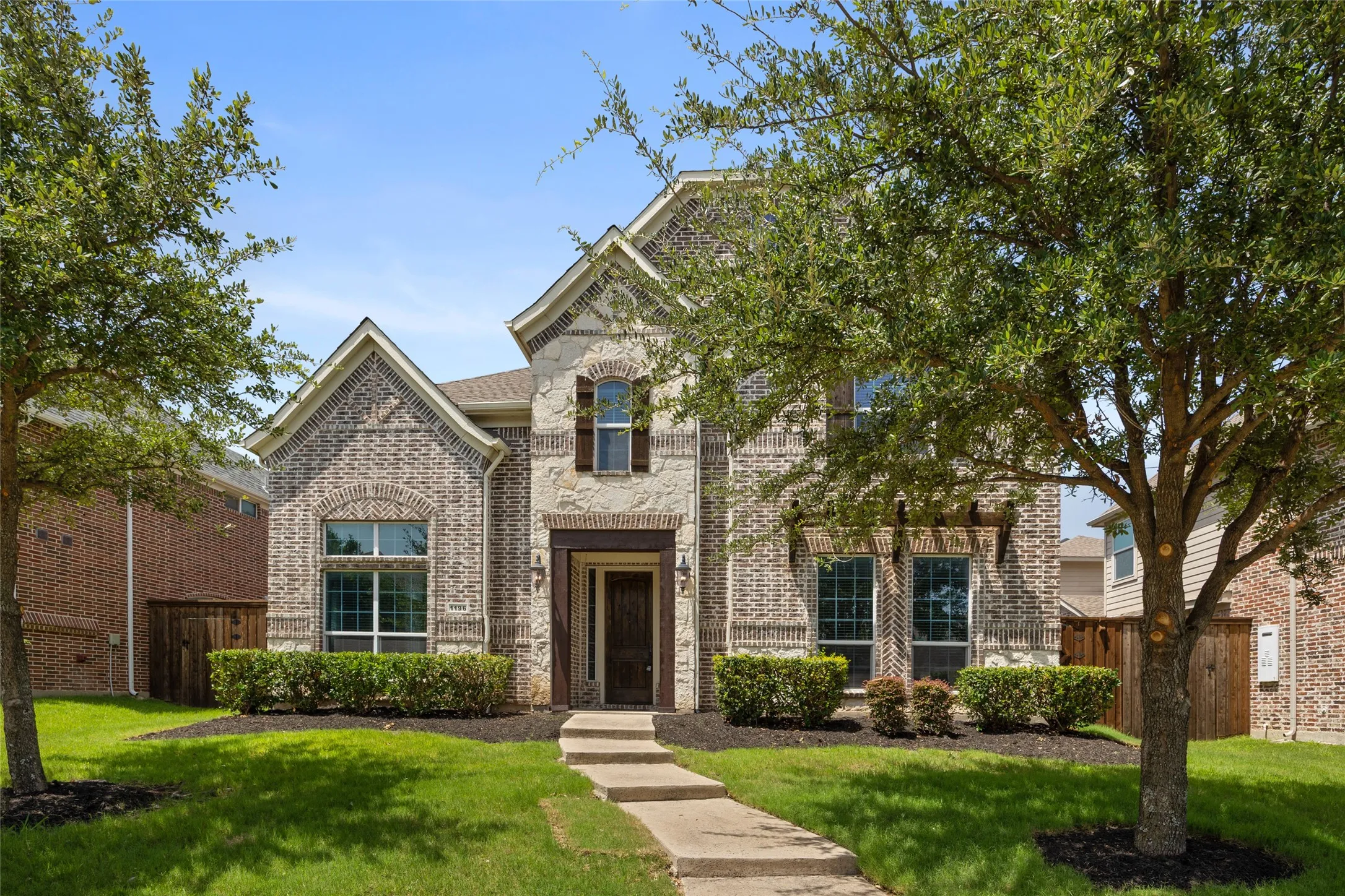 Brick and stone facade residence featuring mature trees, manicured landscaping, a paved walkway, and decorative exterior shutters