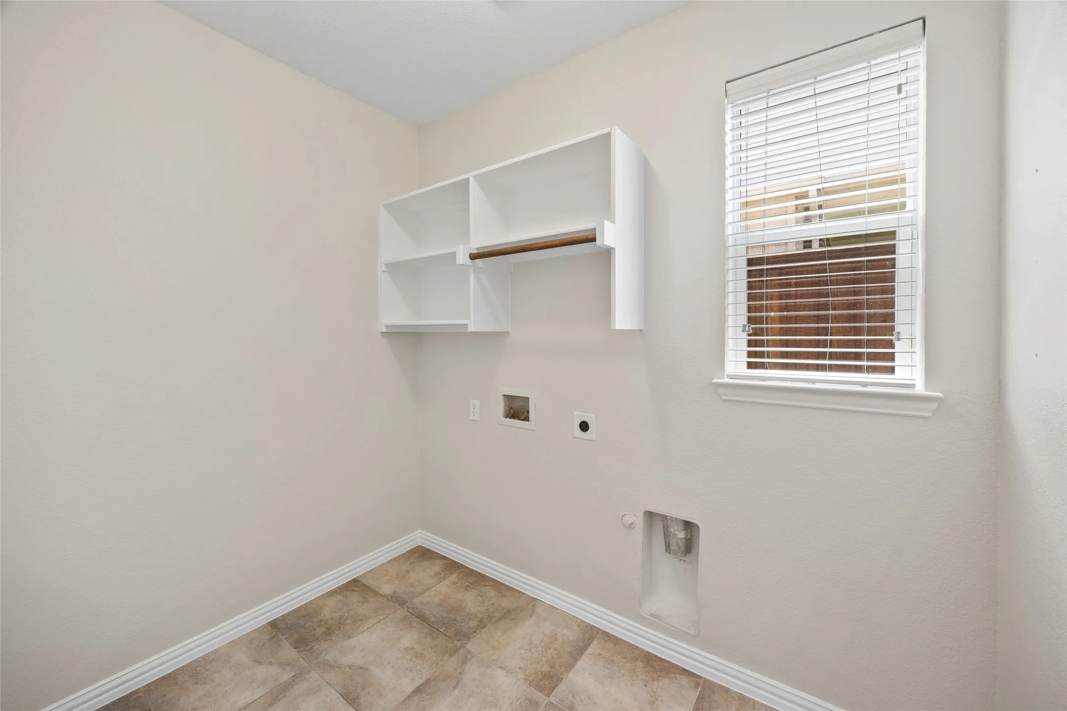 Dedicated utility space featuring tile flooring, white baseboards, a single window with blinds, and built-in upper shelving with a hanging rod