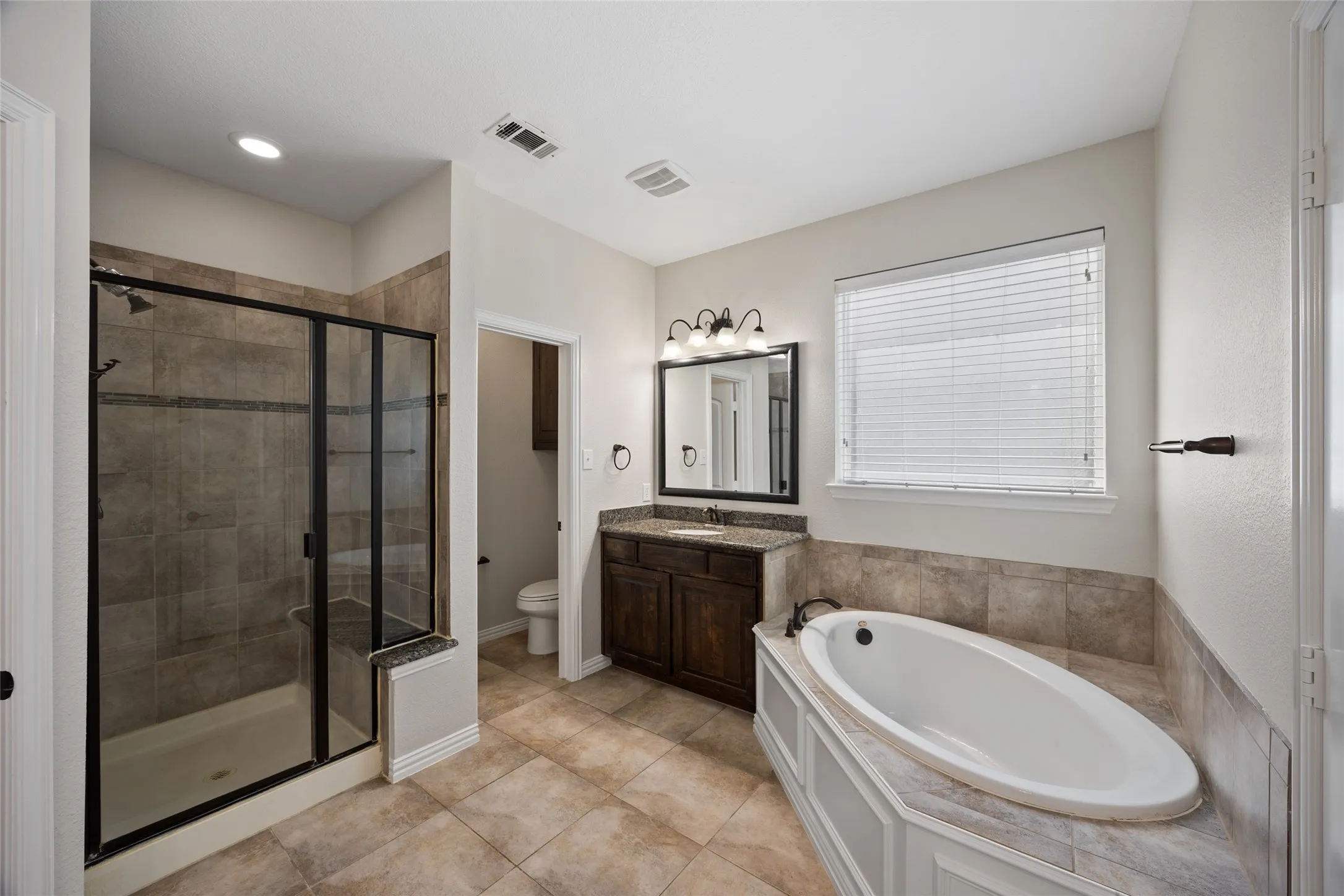 Bathroom featuring a built-in soaking tub, a dark wood vanity with a granite countertop, a framed mirror, an enclosed shower with a built-in bench, and tile flooring