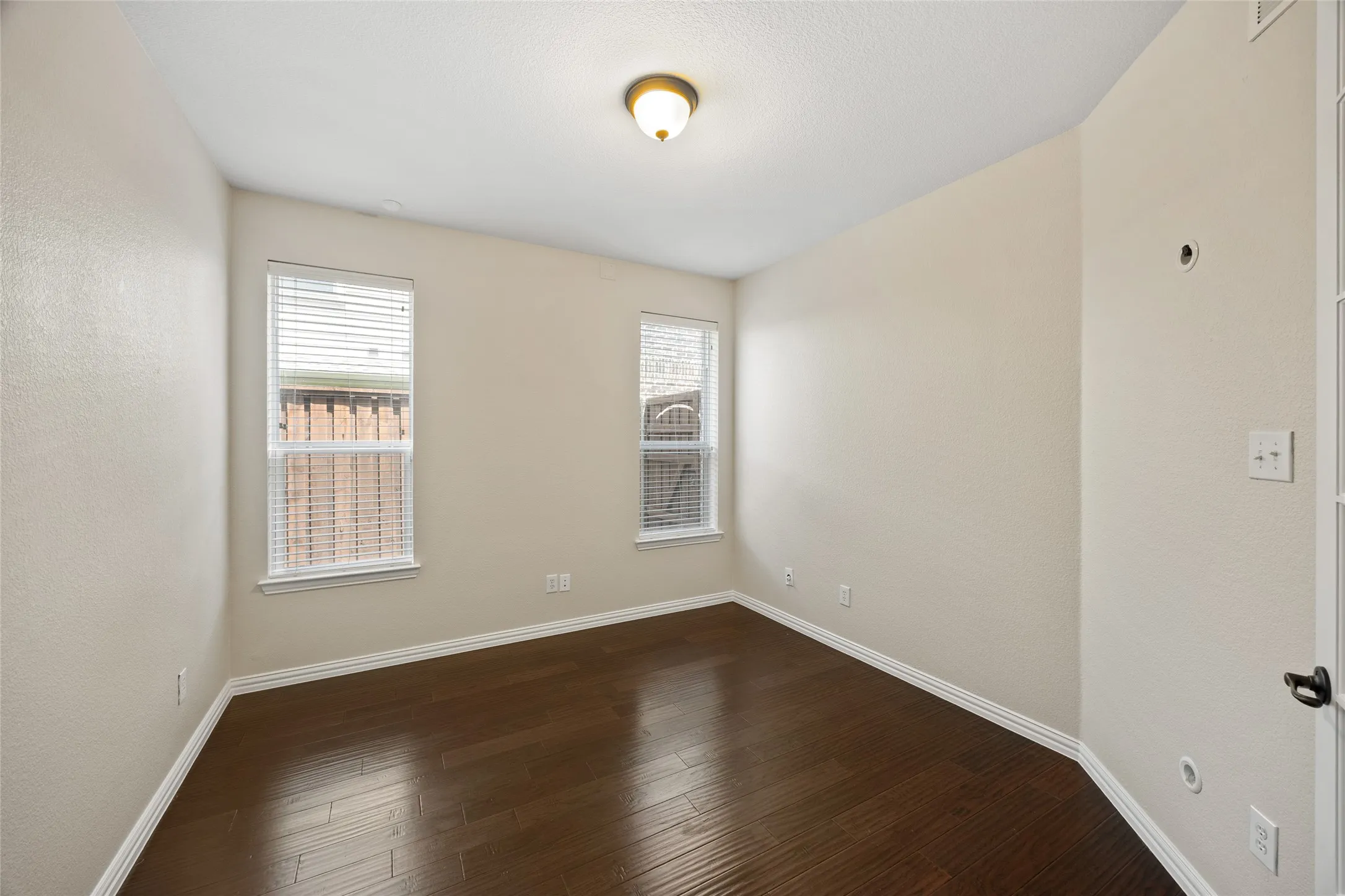 Room featuring dark wood-finish flooring, two windows with blinds, a ceiling-mounted light fixture, and light-toned walls
