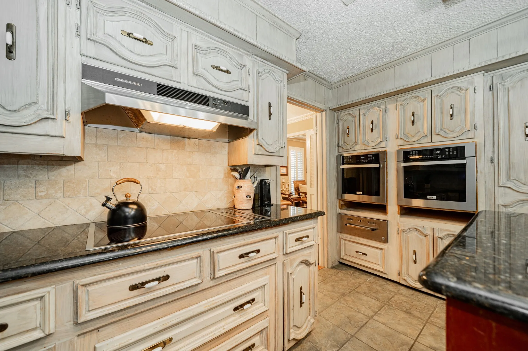 Kitchen featuring tasteful backsplash, under cabinet range hood, a textured ceiling, a warming drawer, and dark stone counters