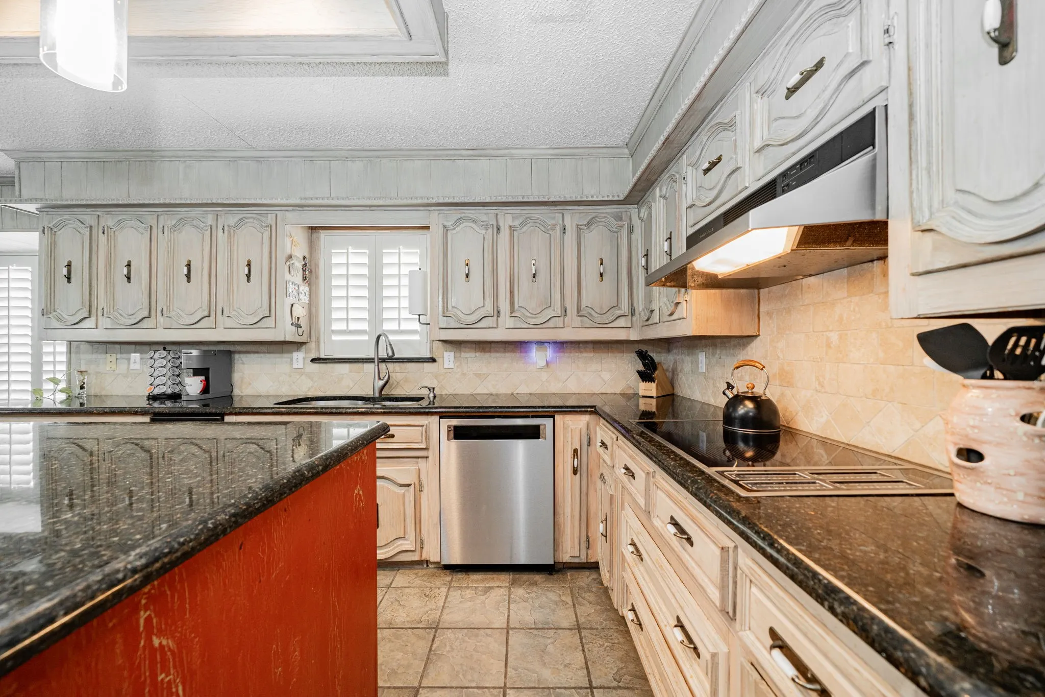Kitchen featuring dark stone countertops, stainless steel dishwasher, backsplash, under cabinet range hood, and a textured ceiling