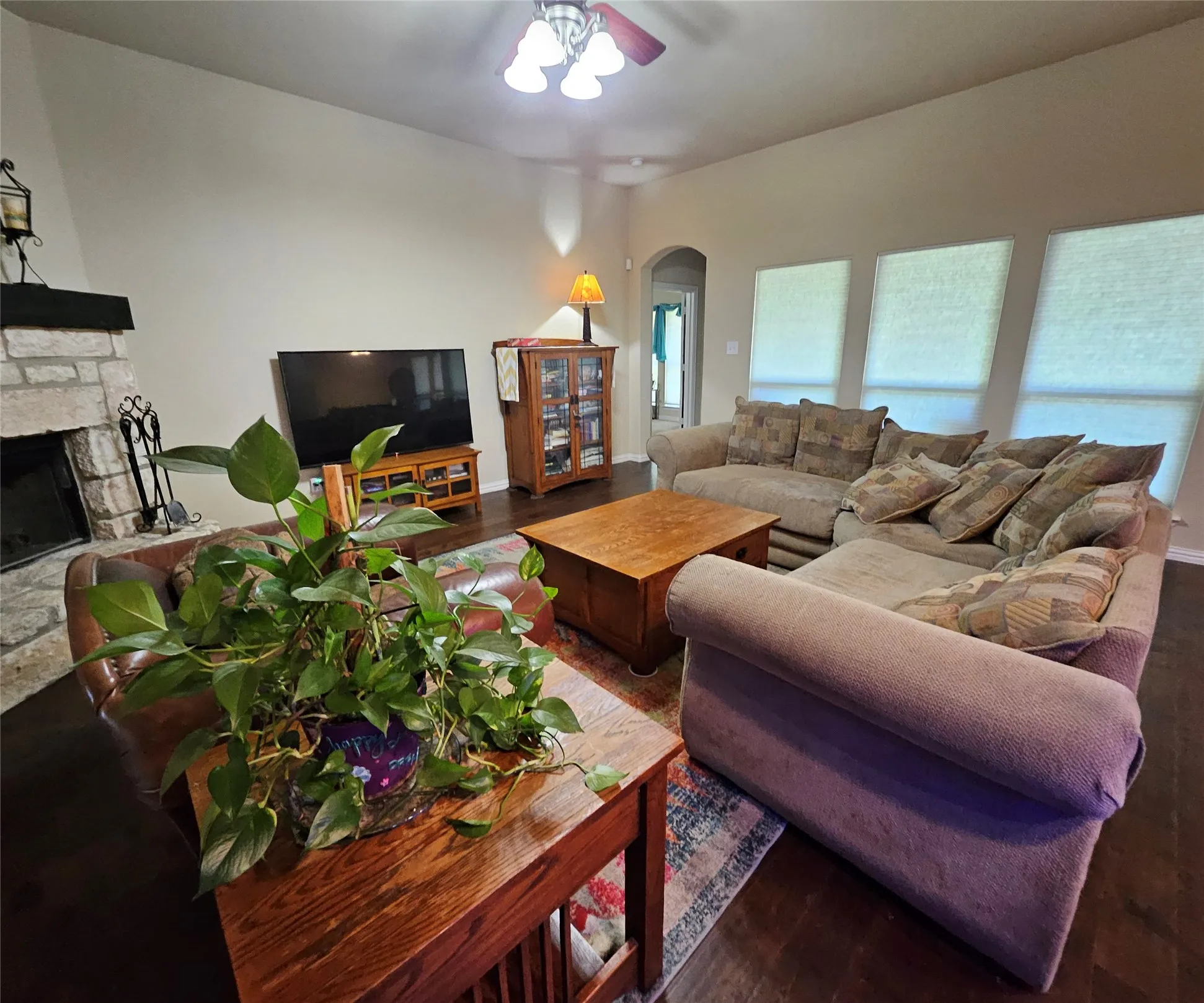 Spacious living area featuring dark wood-finish flooring, a stone fireplace with a dark wood mantel, and multiple windows with blinds