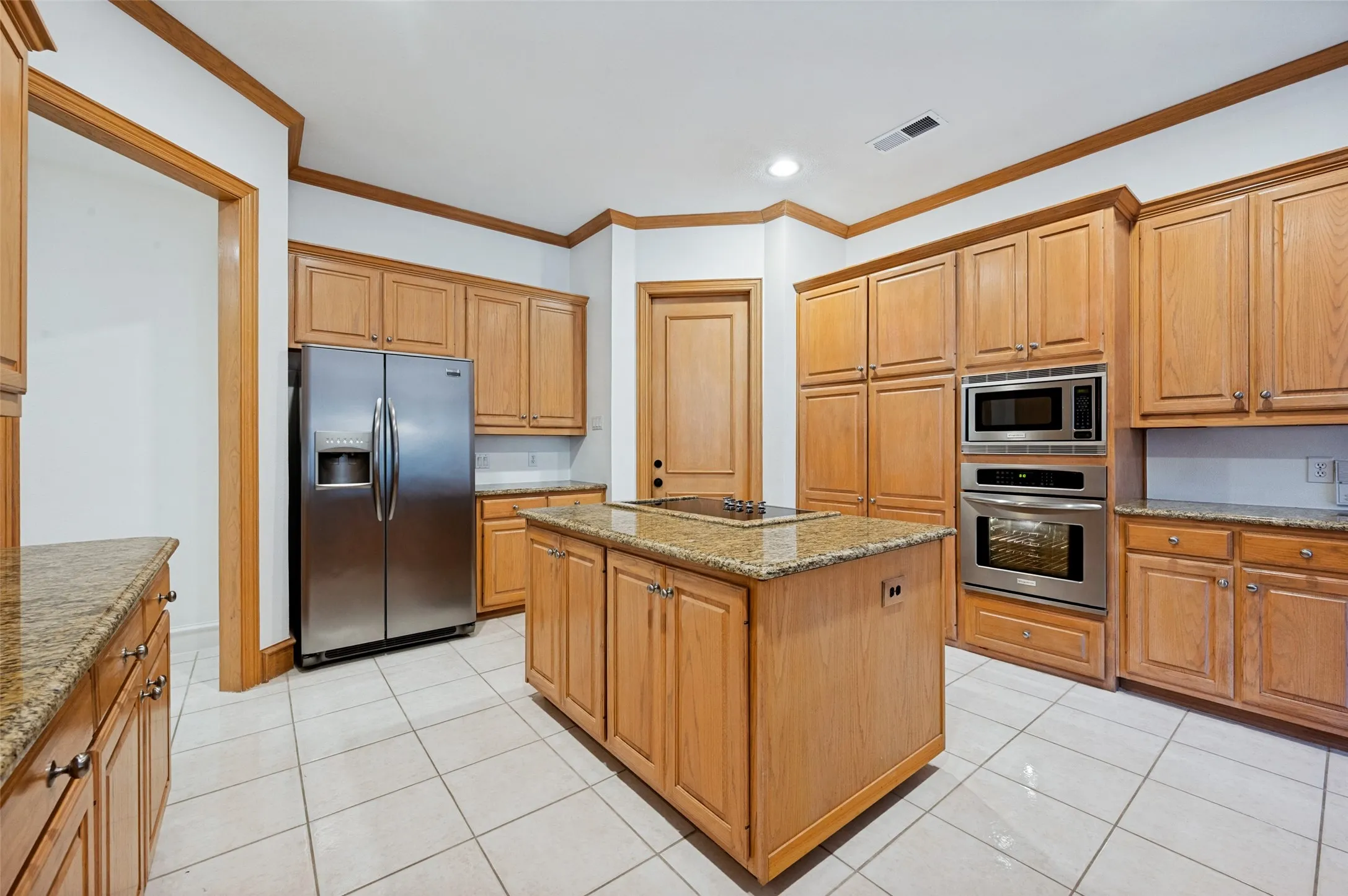 Kitchen featuring a central island with a built-in cooktop, extensive cabinetry with light wood finishes, stainless steel appliances including a double-door refrigerator and double oven, and light-toned tile flooring