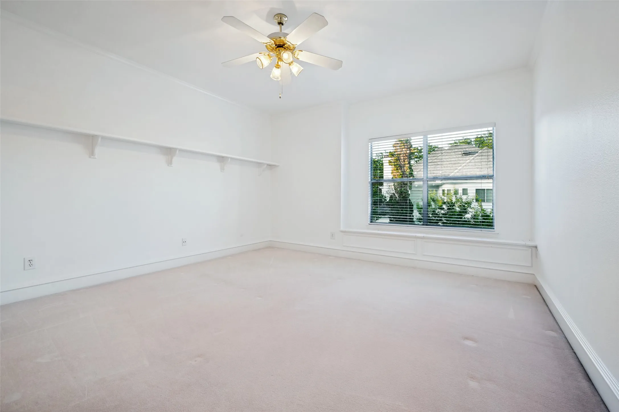 Bright room featuring a large window with blinds, a ceiling fan, light-colored carpeting, white baseboards, and a built-in wall shelf