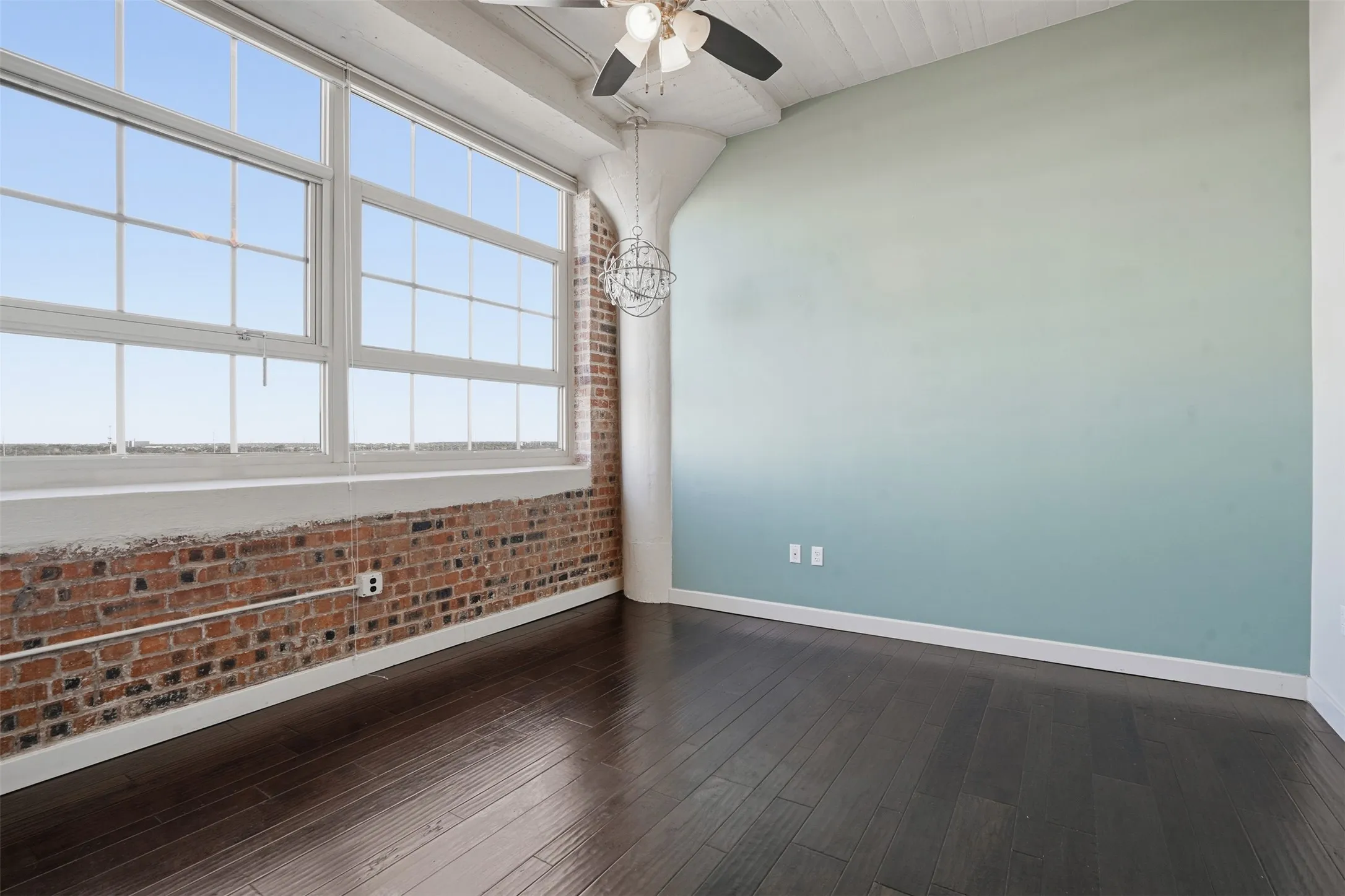 Expansive industrial-style windows, exposed brick wall, dark wood-finish flooring, architectural ceiling fan, and sloped white ceiling